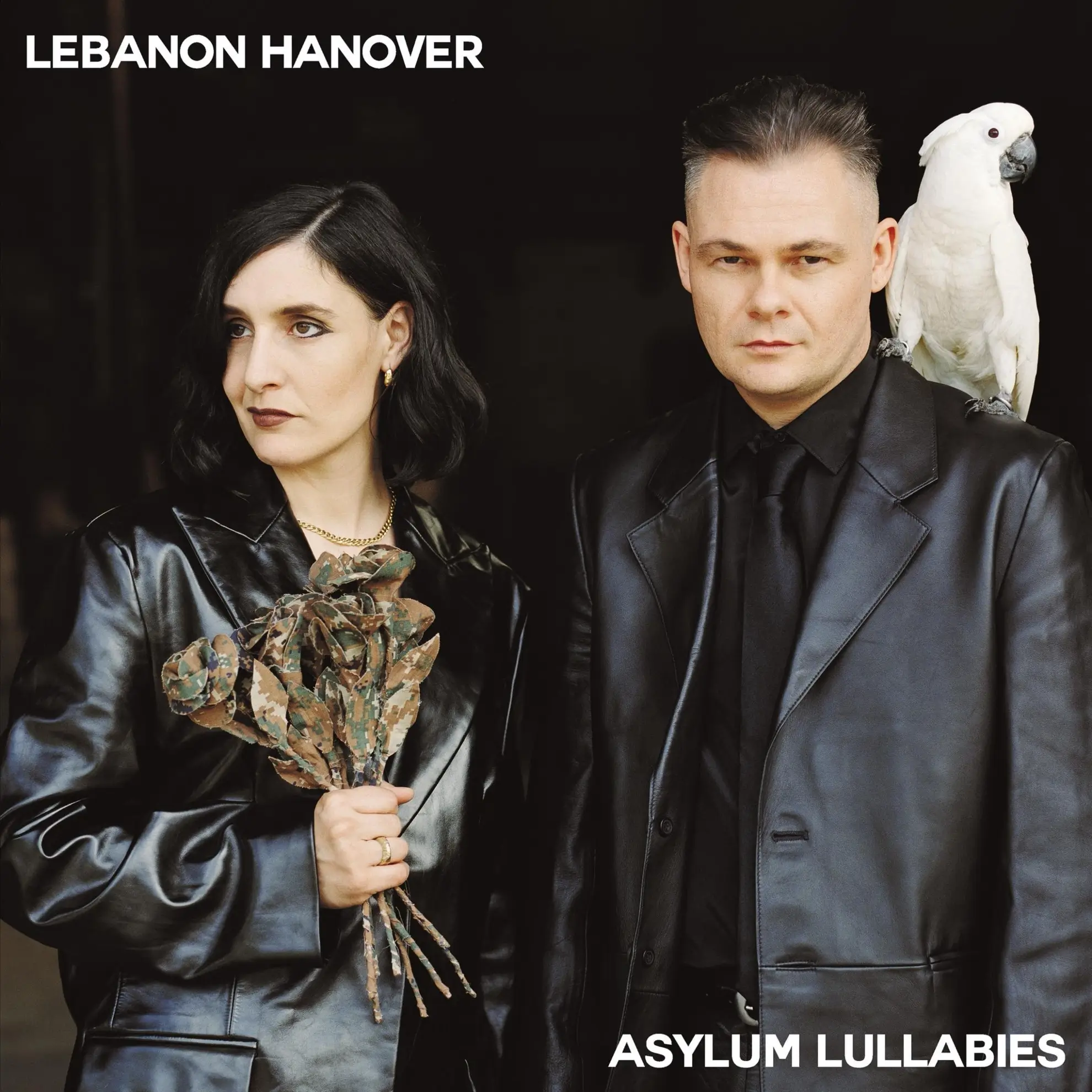 For the ‘Asylum Lullabies’ cover, Lebanon Hanover poses in black leather. A cockatoo is on his shoulder; she holds dark flowers.