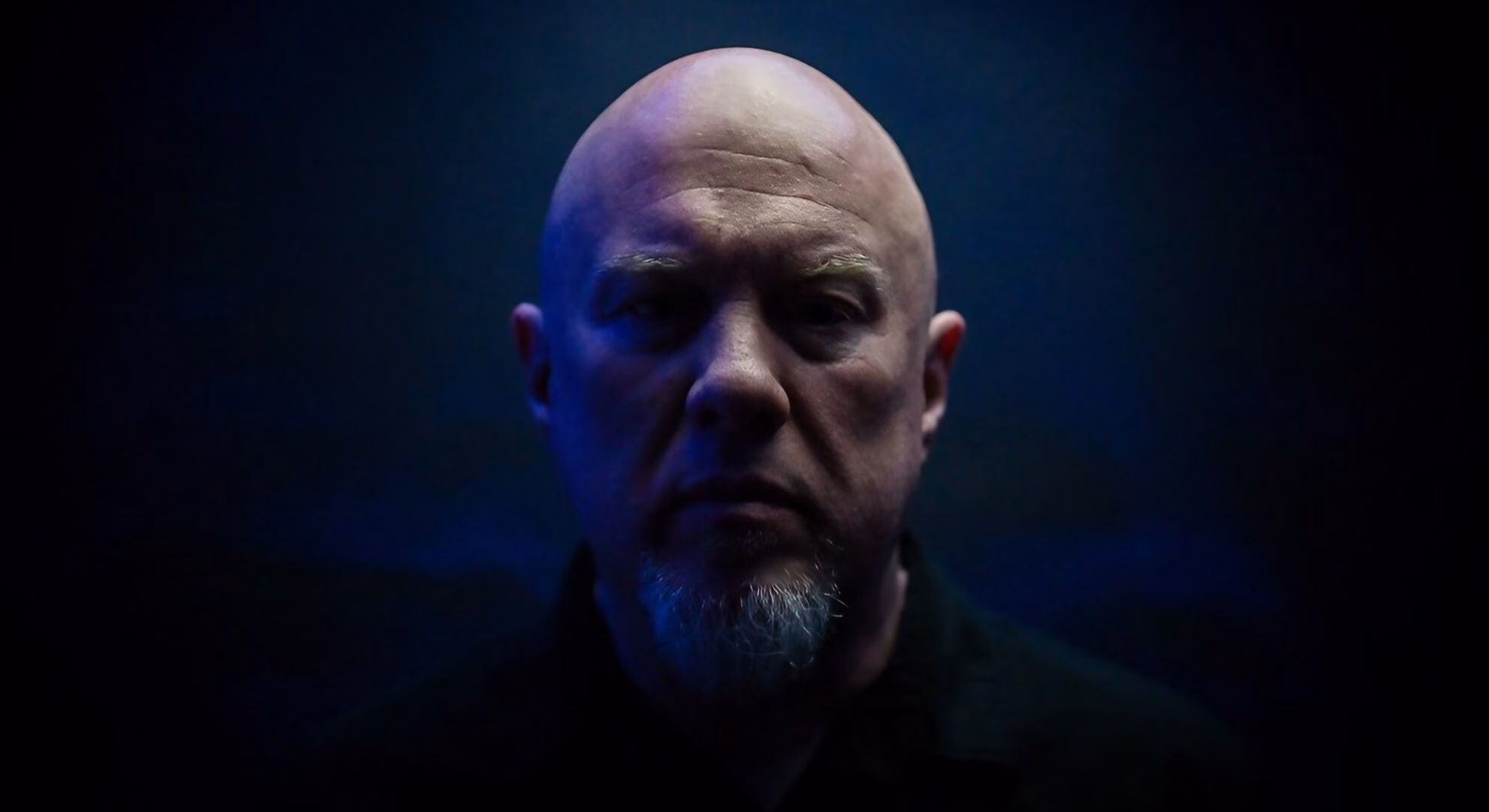 A headshot of Tom Shear of Assemblage 23, looking seriously at the camera in low, moody lighting with a blue hue.