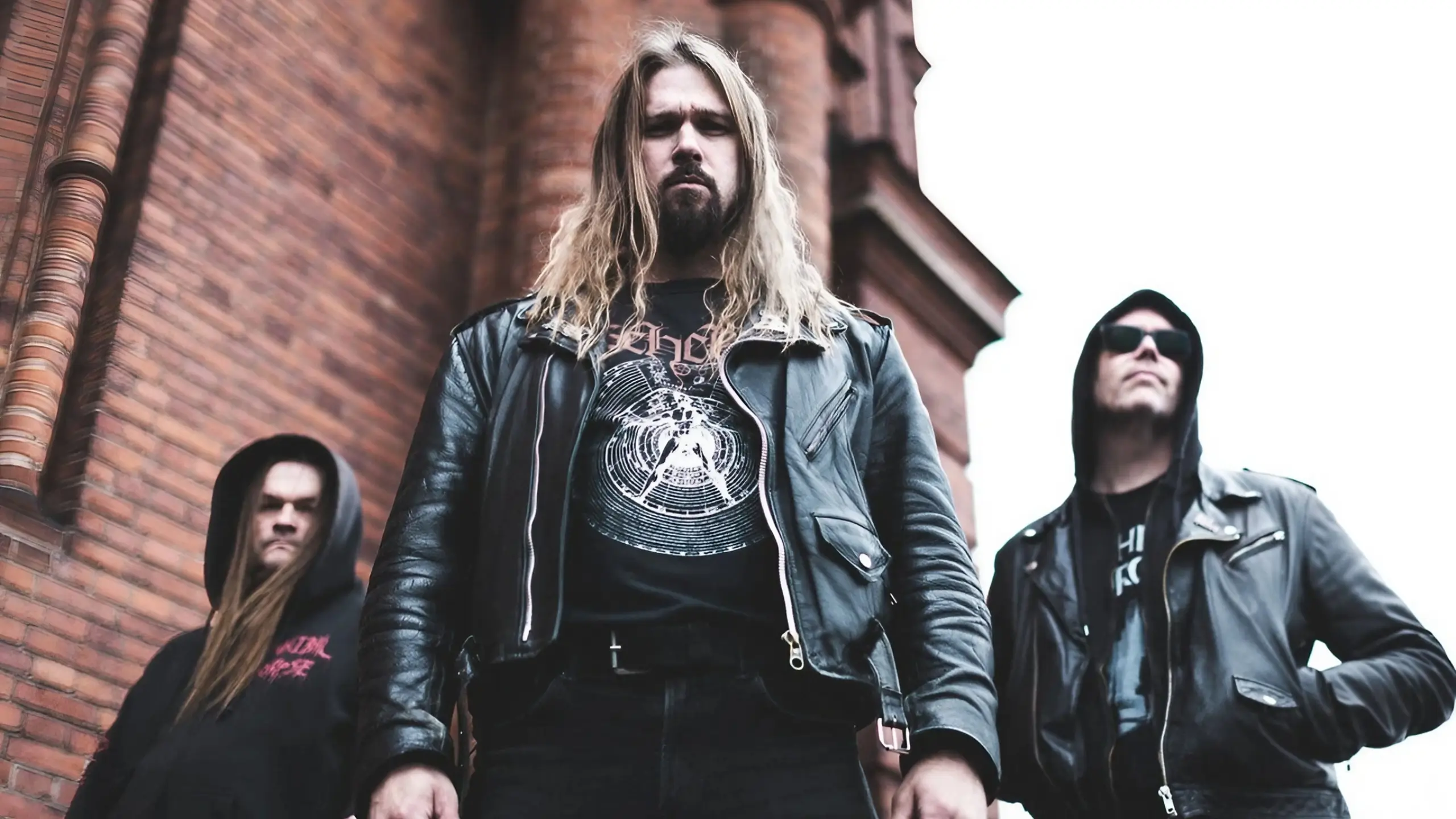 A low-angle shot of three men in black leather jackets standing before a red brick building under an overcast sky.