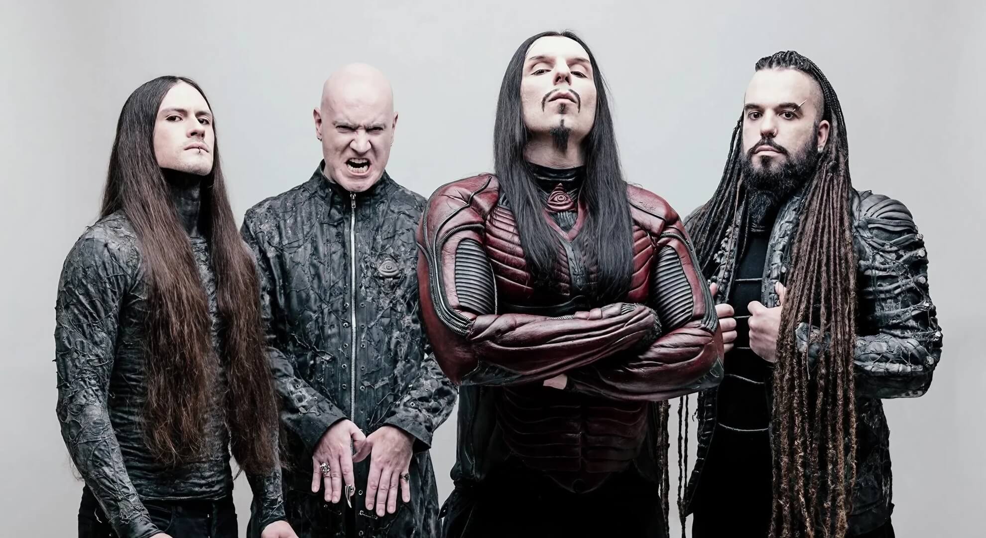 The four members of the band Septicflesh stand together against a light grey background. They are dressed in elaborate, dark, textured, futuristic costumes. The central figure has his arms crossed, while the man to his left snarls aggressively.
