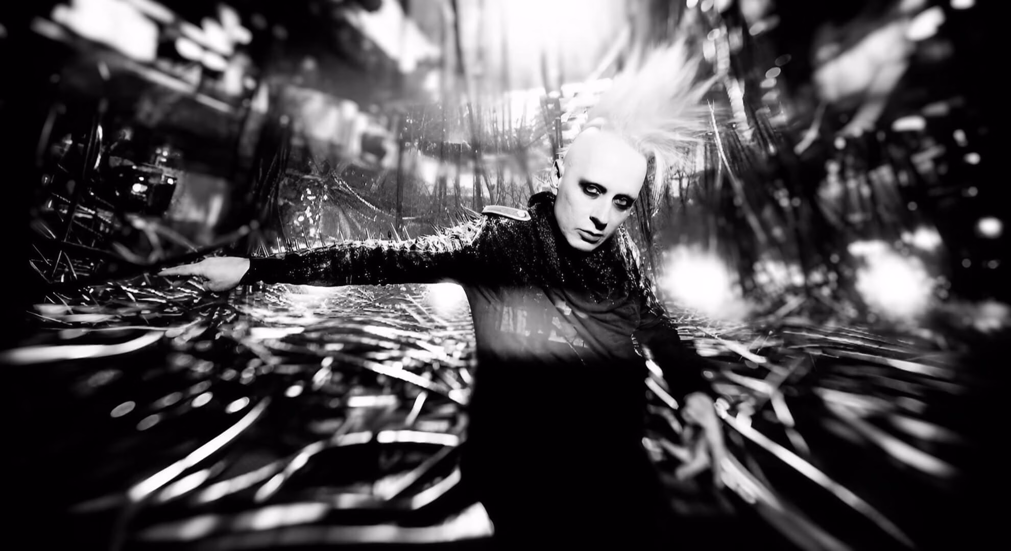 Skold: In the Gleam of the Digital Dollar, an Industrial Veteran Finds His Voice Anew A high-contrast, black-and-white photo of the musician Tim Skold, with his arms outstretched against an abstract, chaotic background.