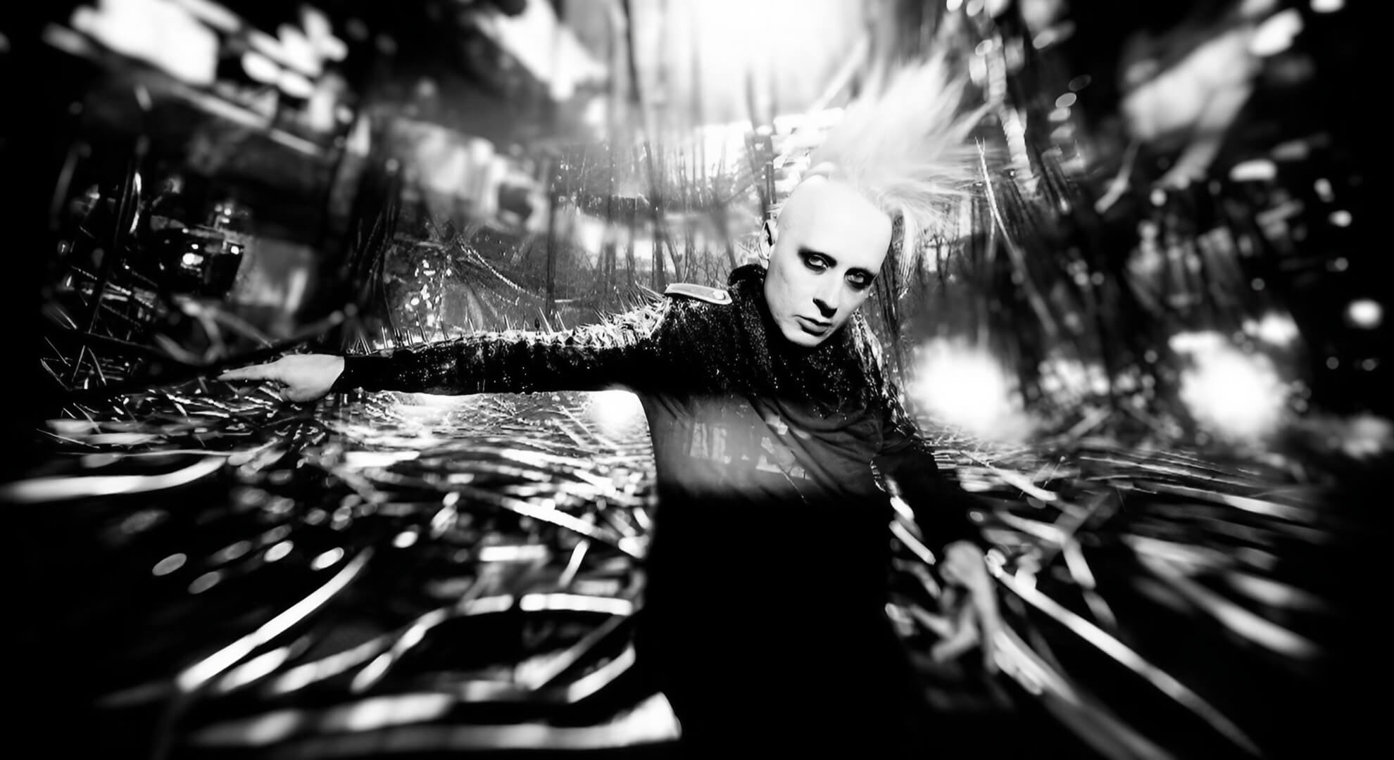 A high-contrast, black-and-white photo of the musician Tim Skold, with his arms outstretched against an abstract, chaotic background.