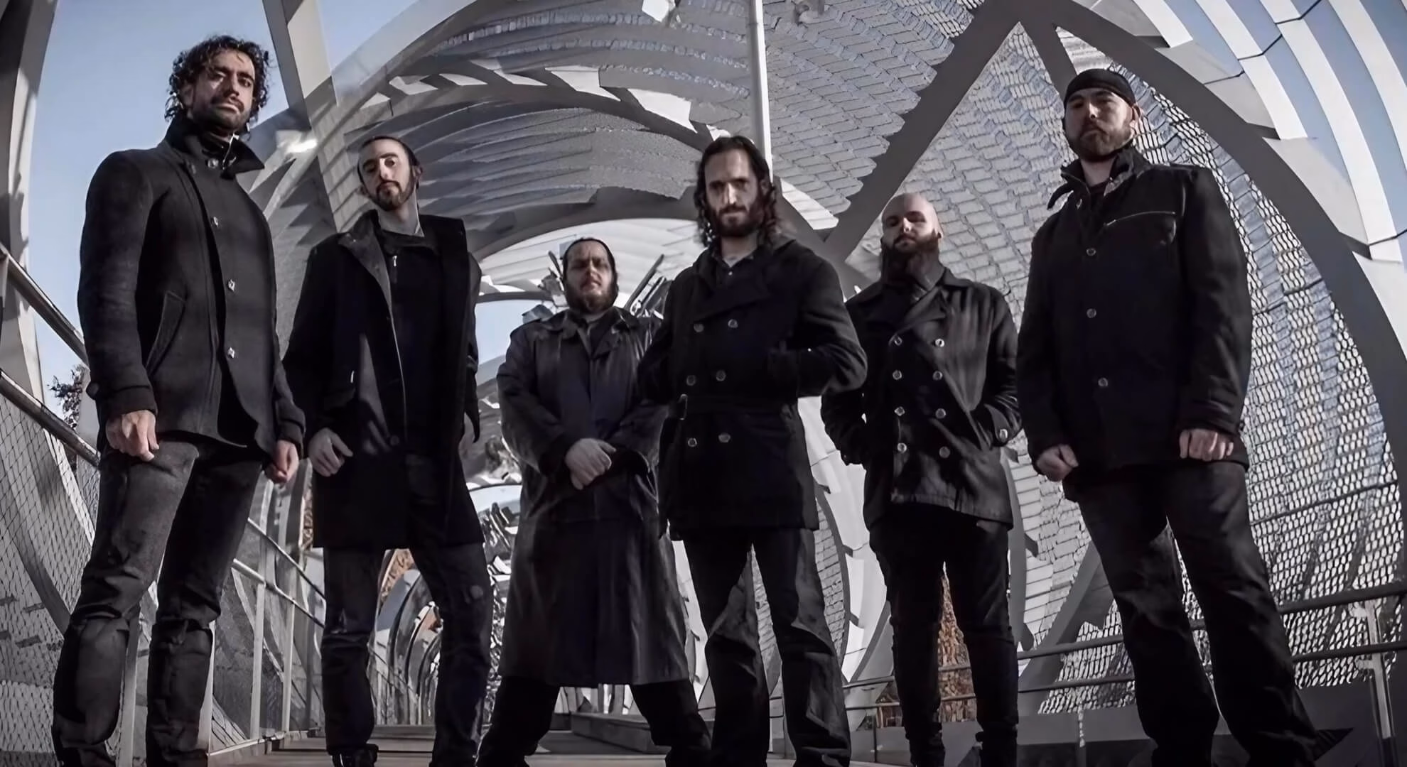 A low-angle shot of the six members of Sun of the Dying standing on a modern architectural bridge. They are all dressed in dark coats and jackets, looking down towards the camera against a backdrop of a complex, swirling metallic structure.
