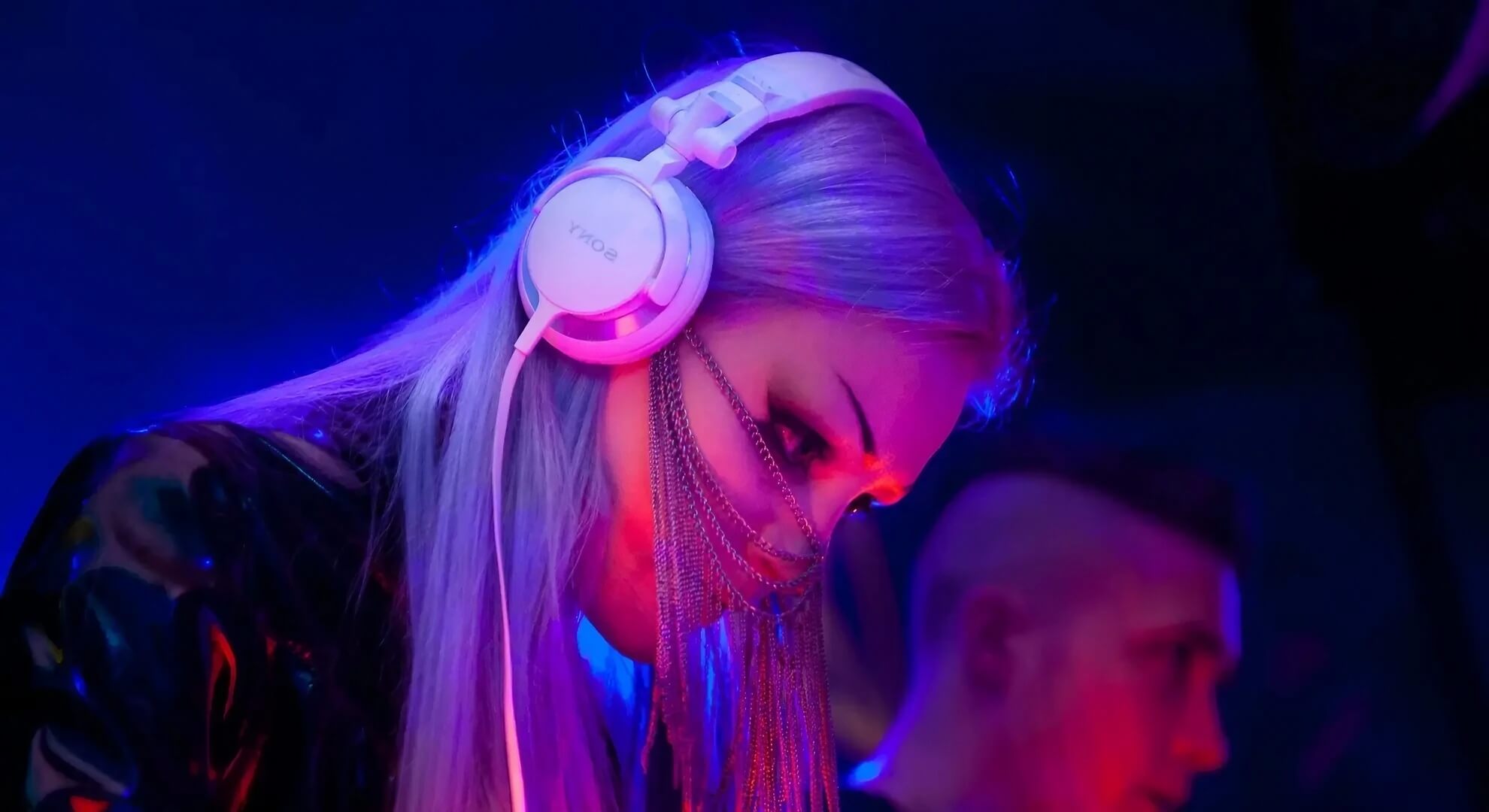 A DJ with long purple hair, wearing white headphones and a silver chain mask, performs in a dark club with blue and purple lighting.