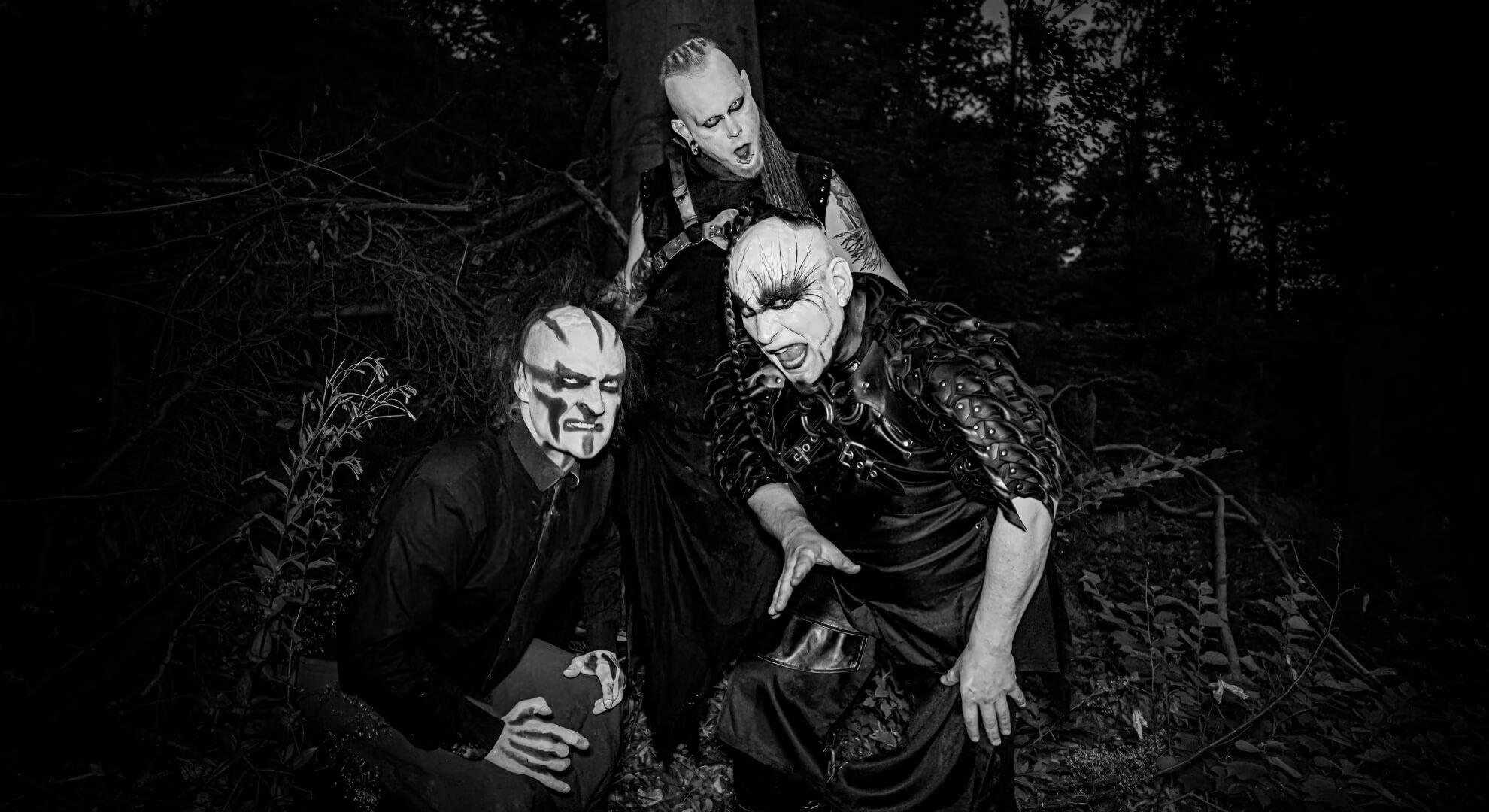 Three men in theatrical makeup and dark clothing pose in a dark, wooded setting.