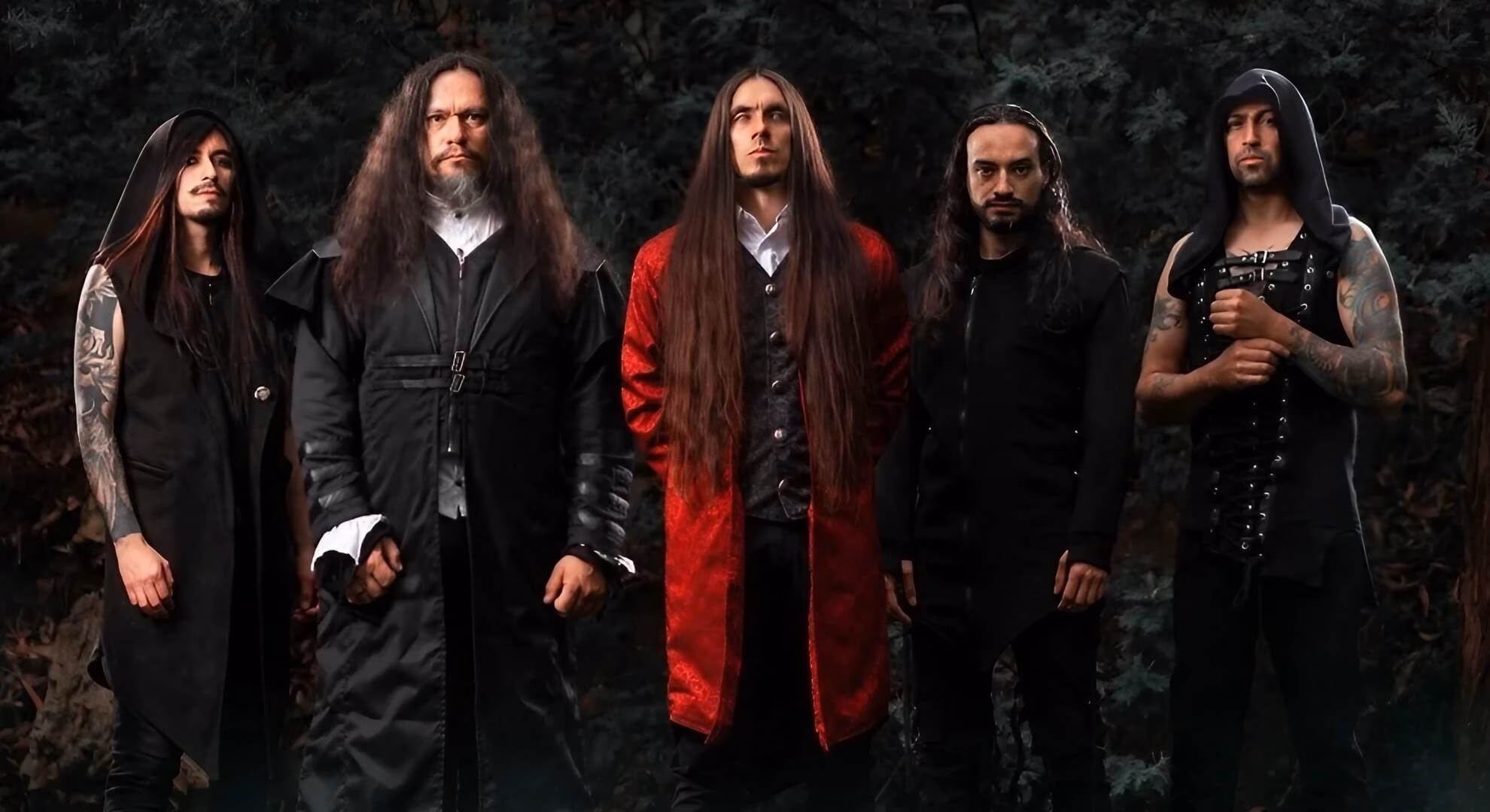 Five men in ornate, dark clothing pose in a forest. The central figure wears a long, red coat over a white shirt.