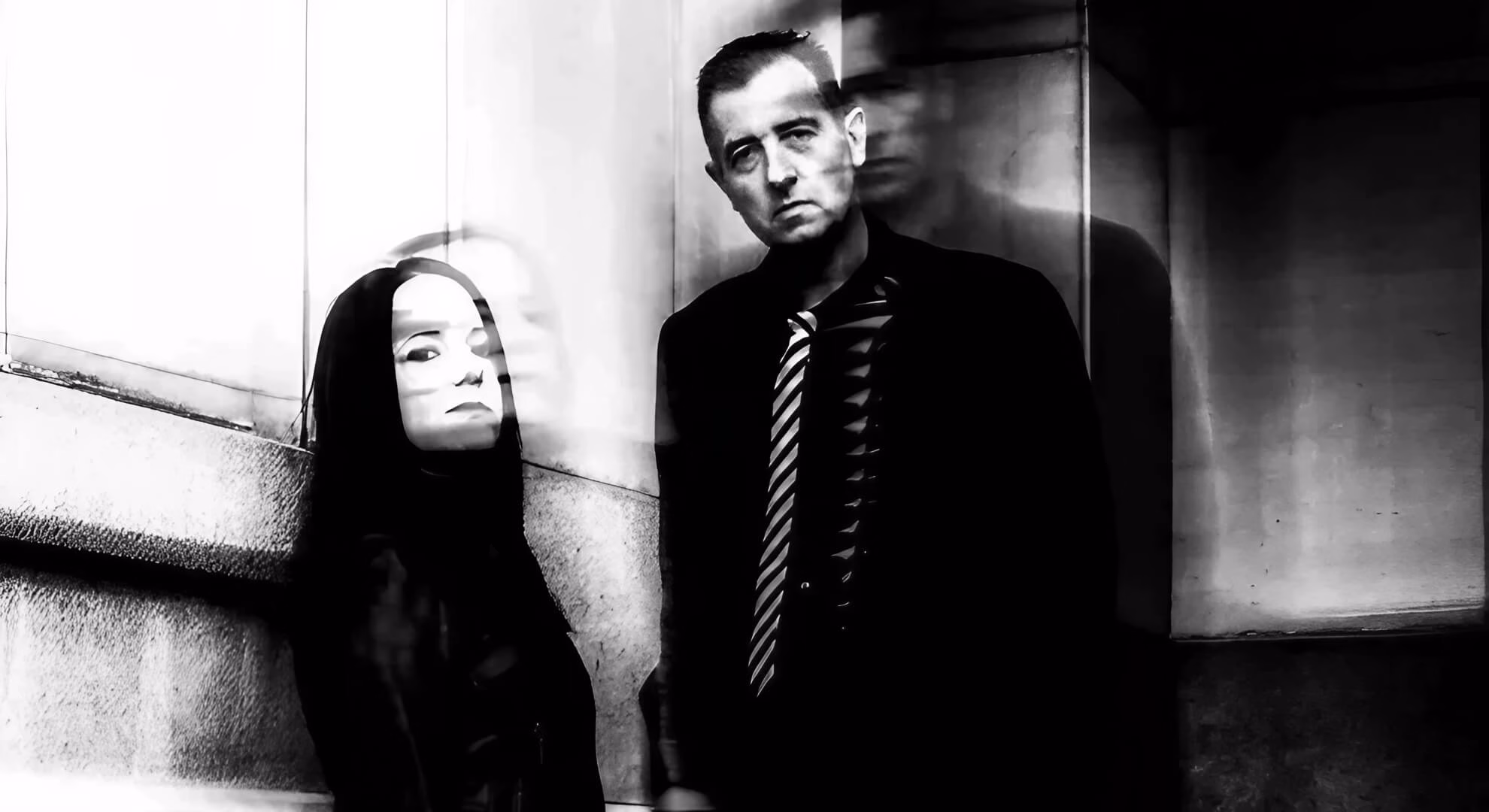 A woman and a man in a suit pose against a wall in a stark, black and white photograph with motion blur.
