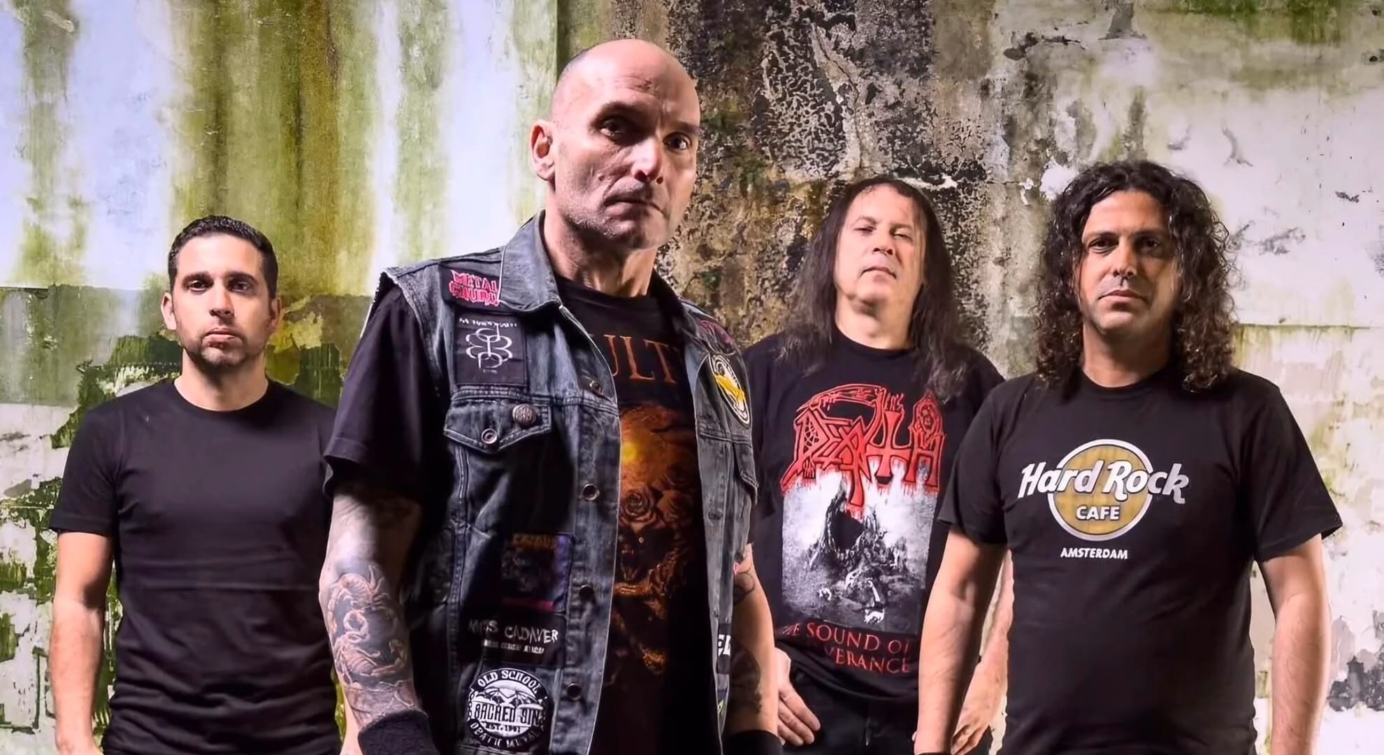 Four men in a metal band pose against a gritty, stained wall. The central figure wears a patched denim vest.