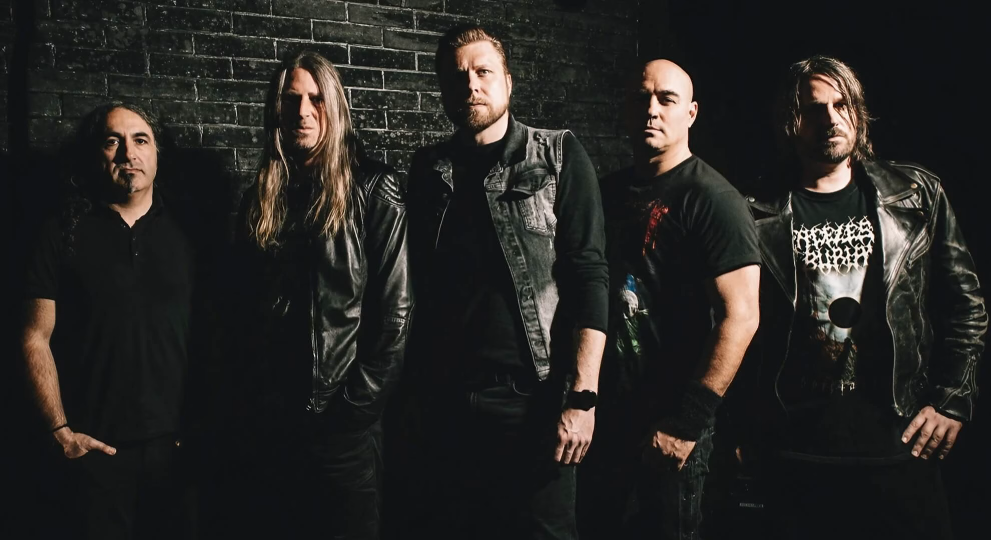 The five members of Night In Gales stand in a line against a dark brick wall, dressed in black leather and denim.