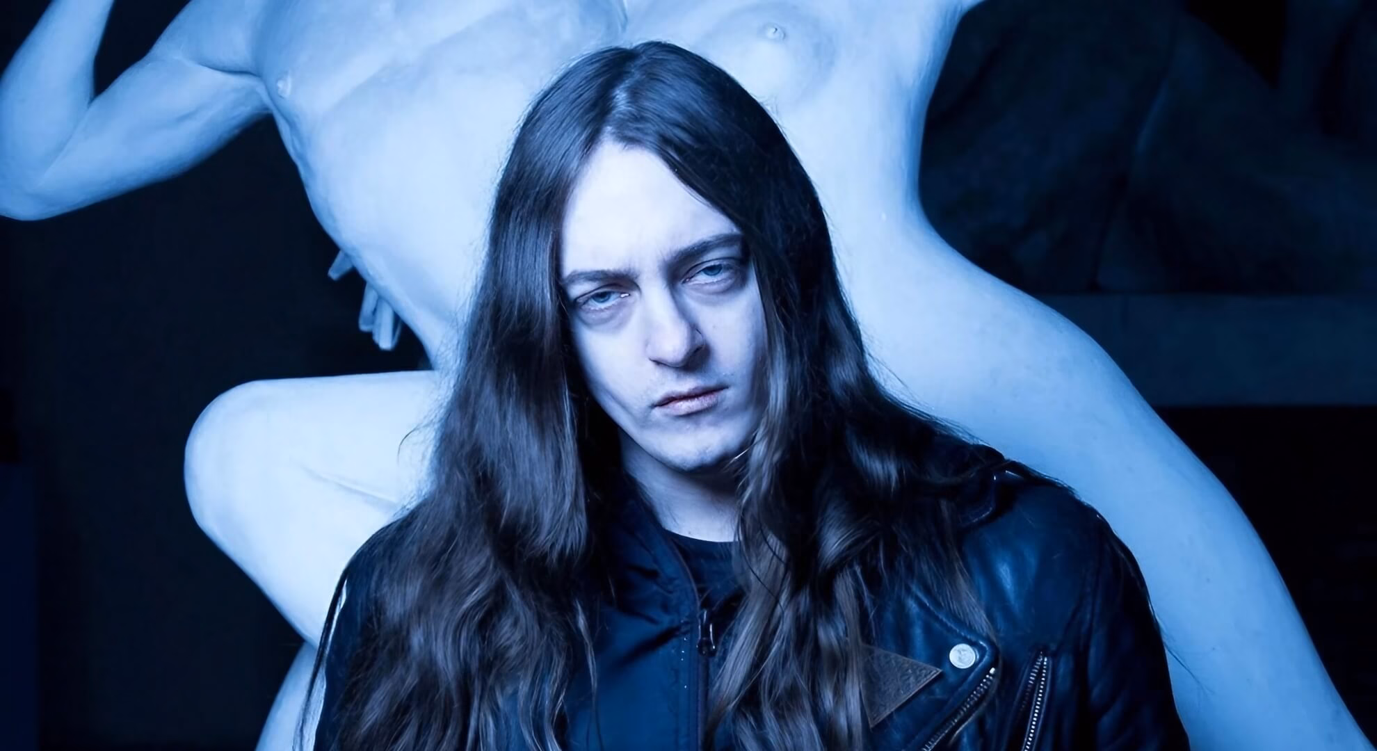 A man with long dark hair and a leather jacket stares intently, illuminated by a cool blue light in front of a pale sculpture.