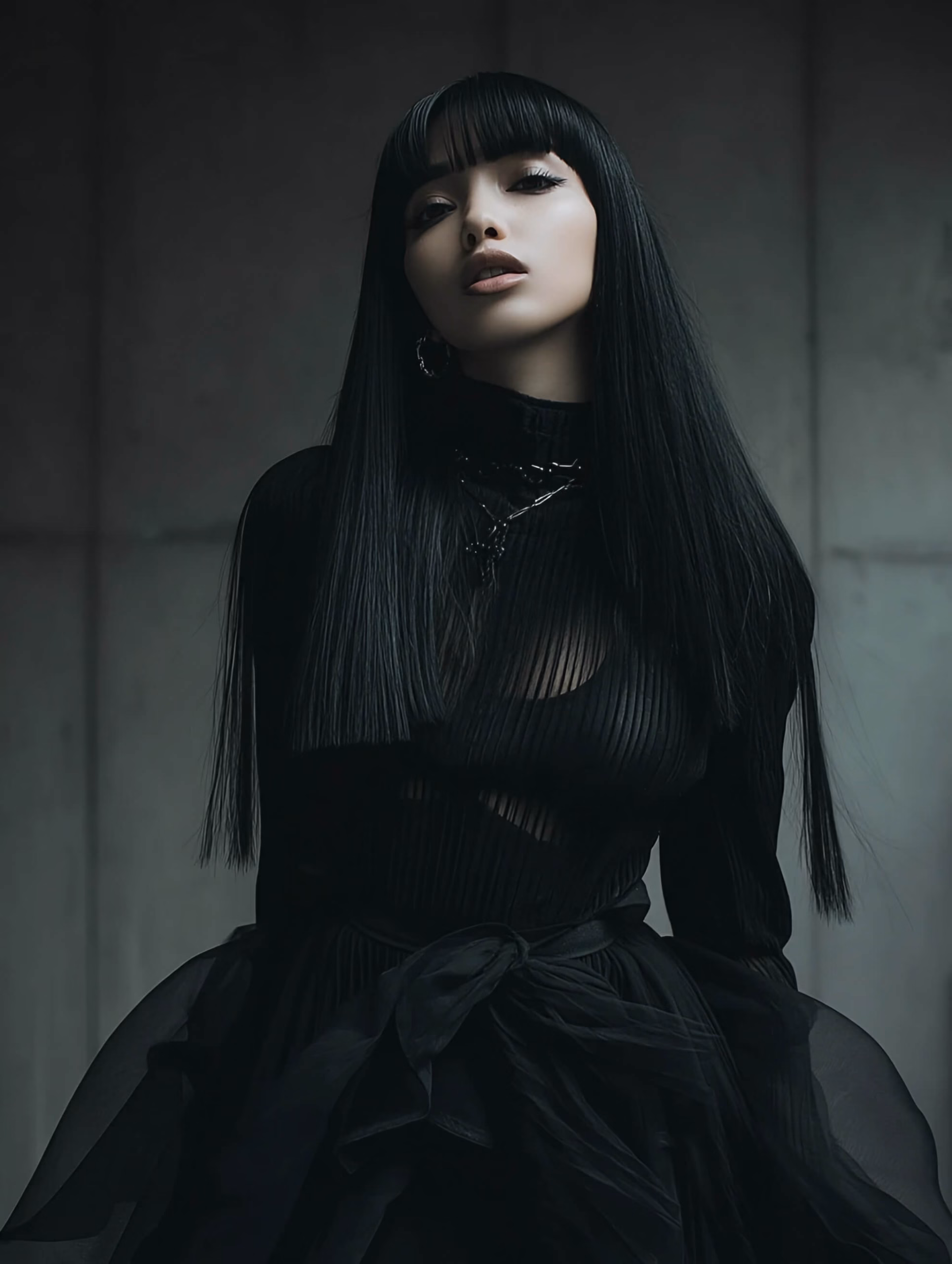 AExpo A woman with long black hair and bangs, wearing a form-fitting black top and a voluminous black skirt, posing in a high-fashion style against a plain gray wall.