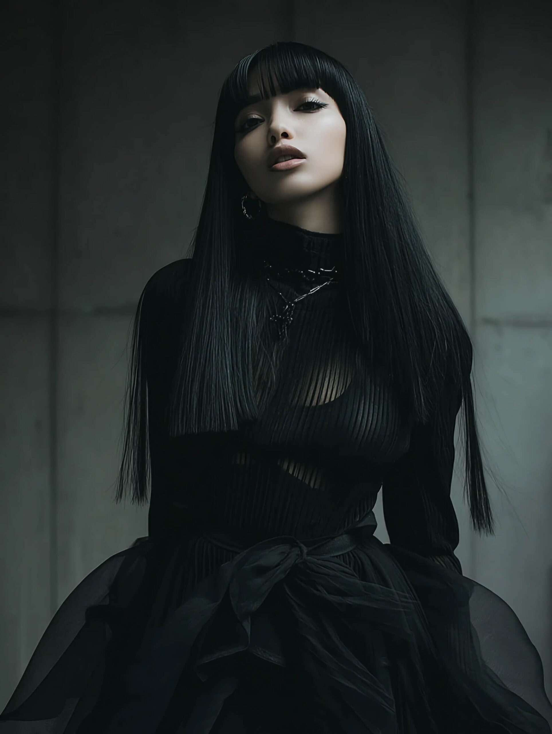 A woman with long black hair and bangs, wearing a form-fitting black top and a voluminous black skirt, posing in a high-fashion style against a plain gray wall.