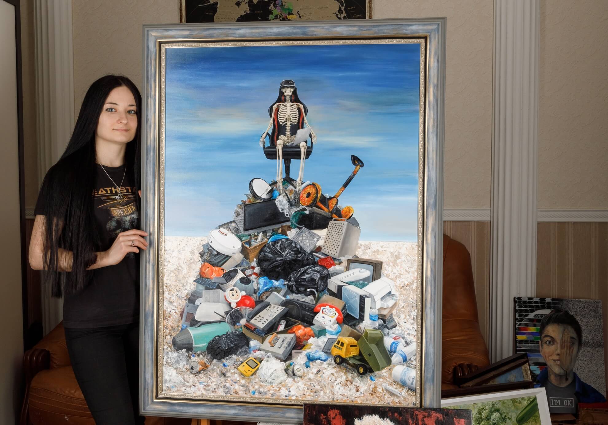 Artist Ellaya Yefymova stands next to her large painting ‘The Apotheosis of invisible struggle,’ which depicts a skeleton wearing a VR headset sitting on a pile of electronic waste and toys.