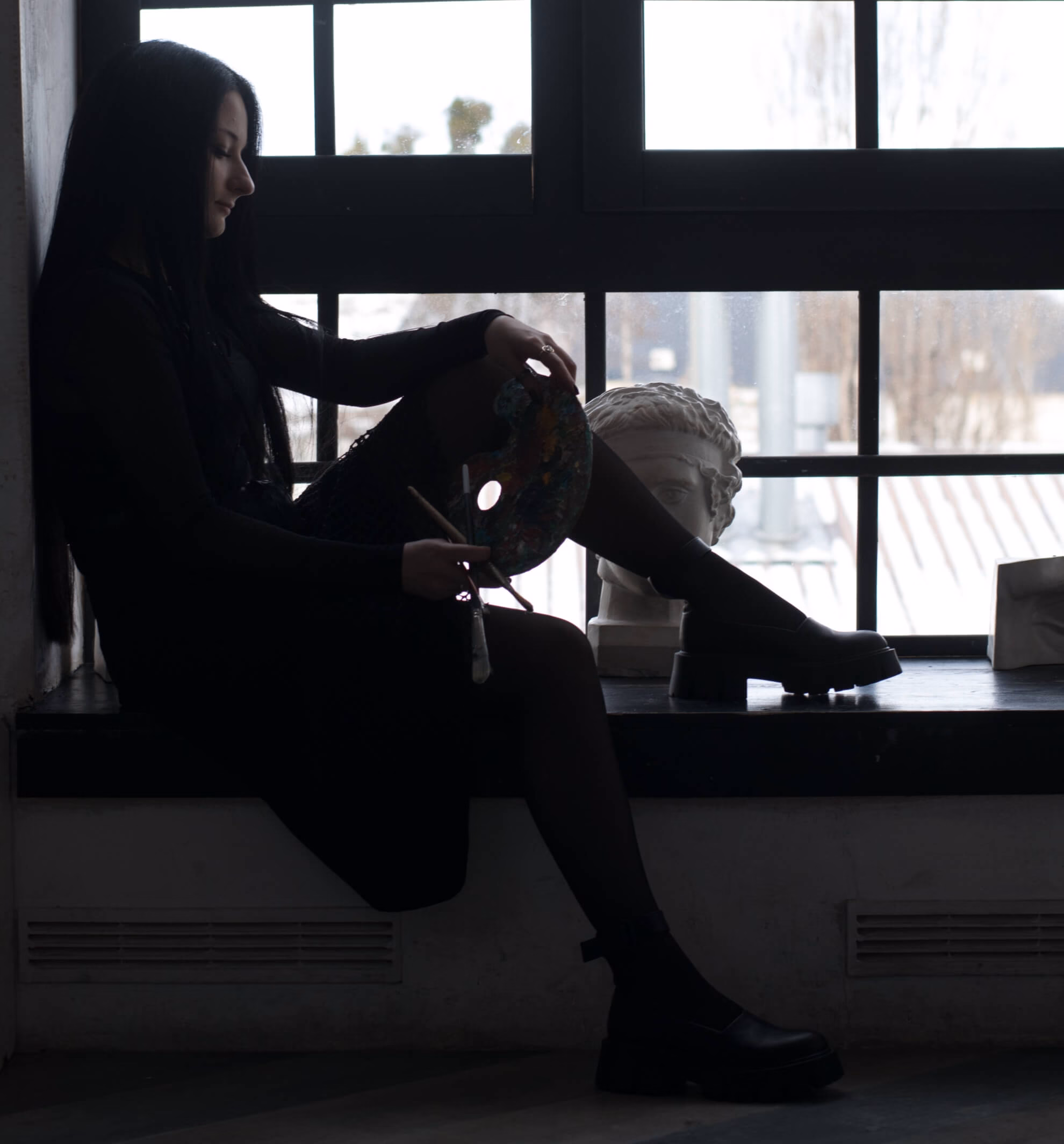 Artist Ellaya Yefymova sits in silhouette on a wide windowsill, holding a paint palette and brushes, with a classical bust sculpture beside her.
