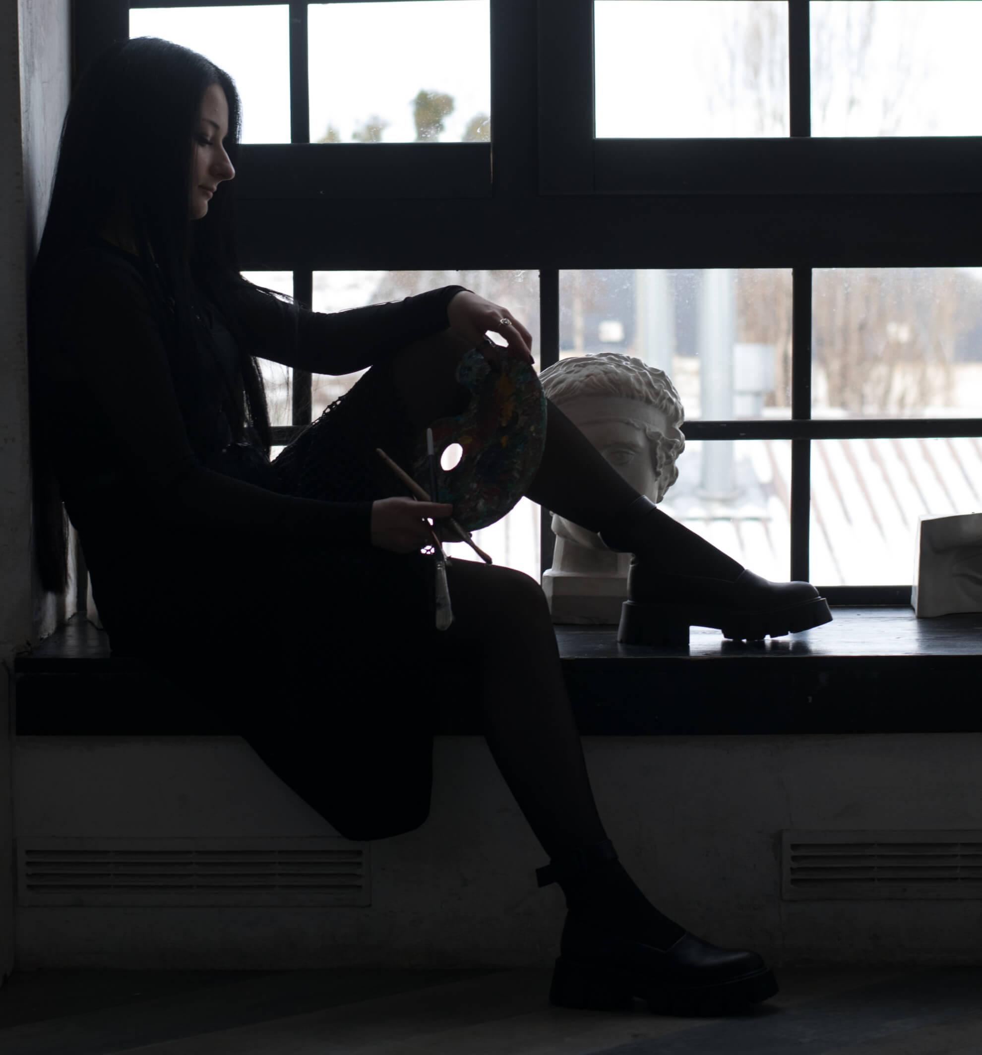 Artist Ellaya Yefymova sits in silhouette on a wide windowsill, holding a paint palette and brushes, with a classical bust sculpture beside her.