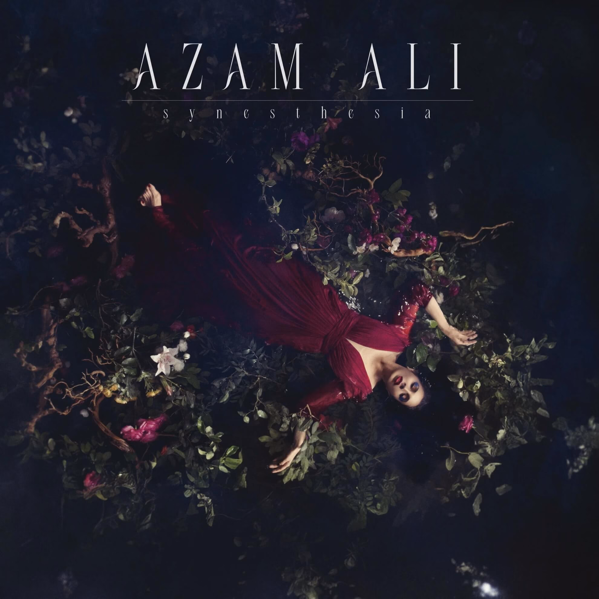 Album cover for &lsquo;Synesthesia.&rsquo; Azam Ali in a red dress floats in dark water surrounded by flowers and leaves.
