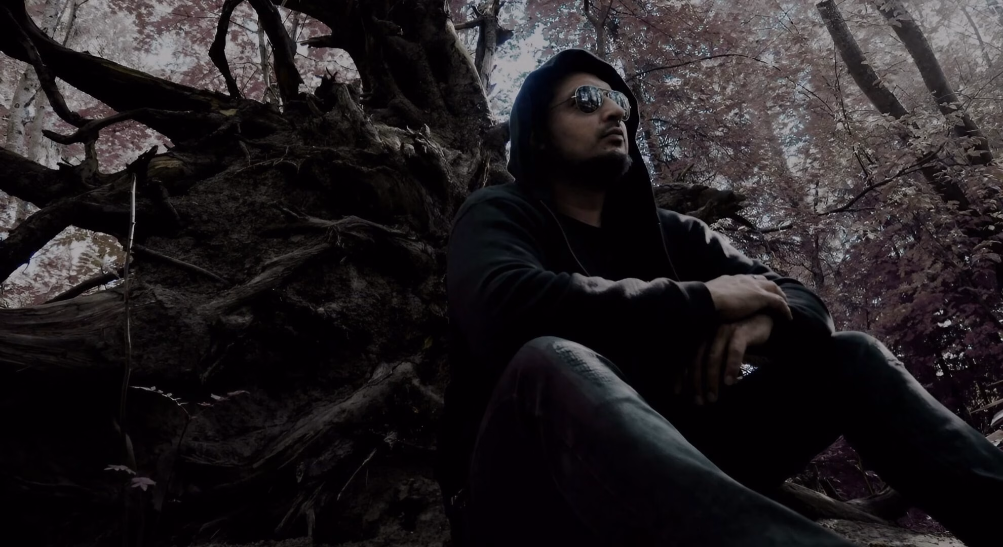 A low-angle shot of a man in a black hoodie and sunglasses sitting against a large, gnarled tree root in a dark forest.