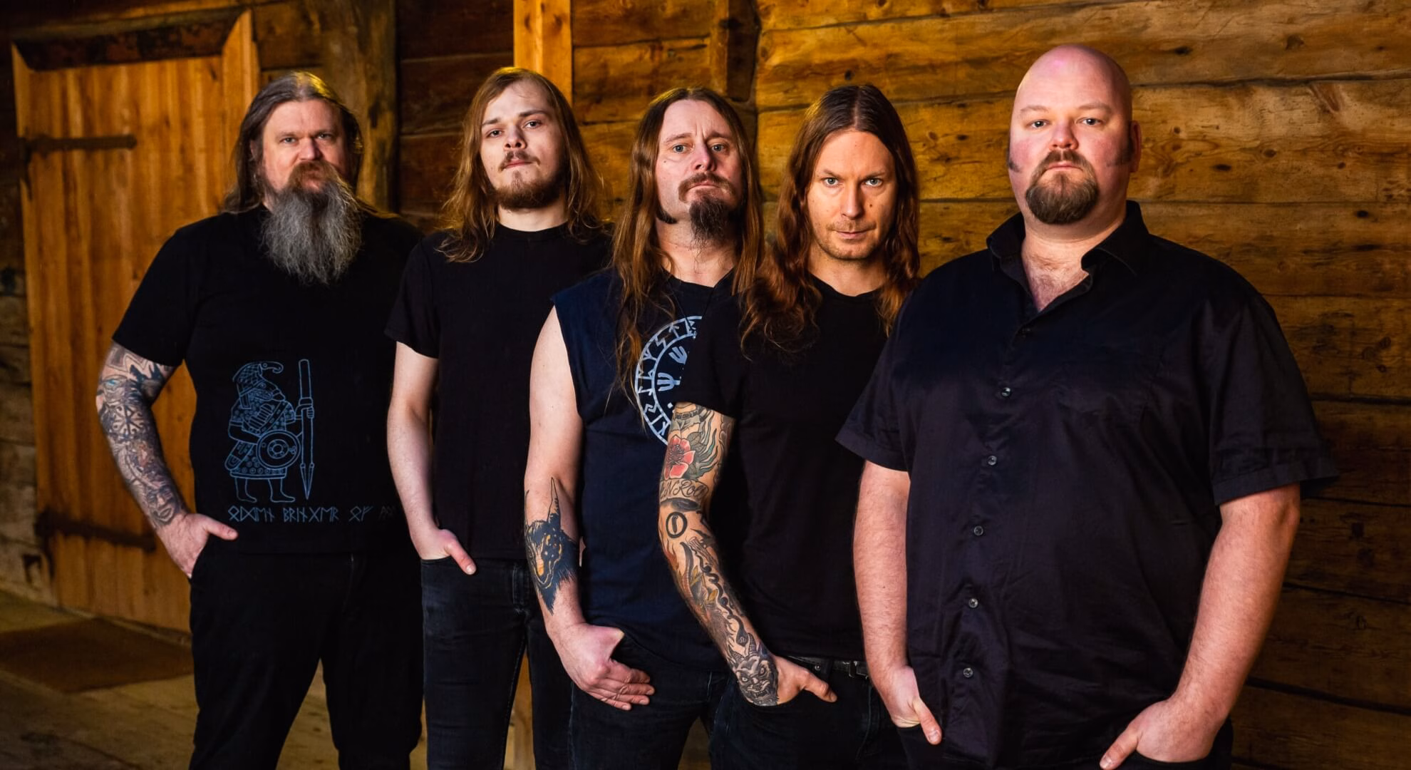 Five men in dark shirts stand in a row against an interior wall of horizontal wooden planks.