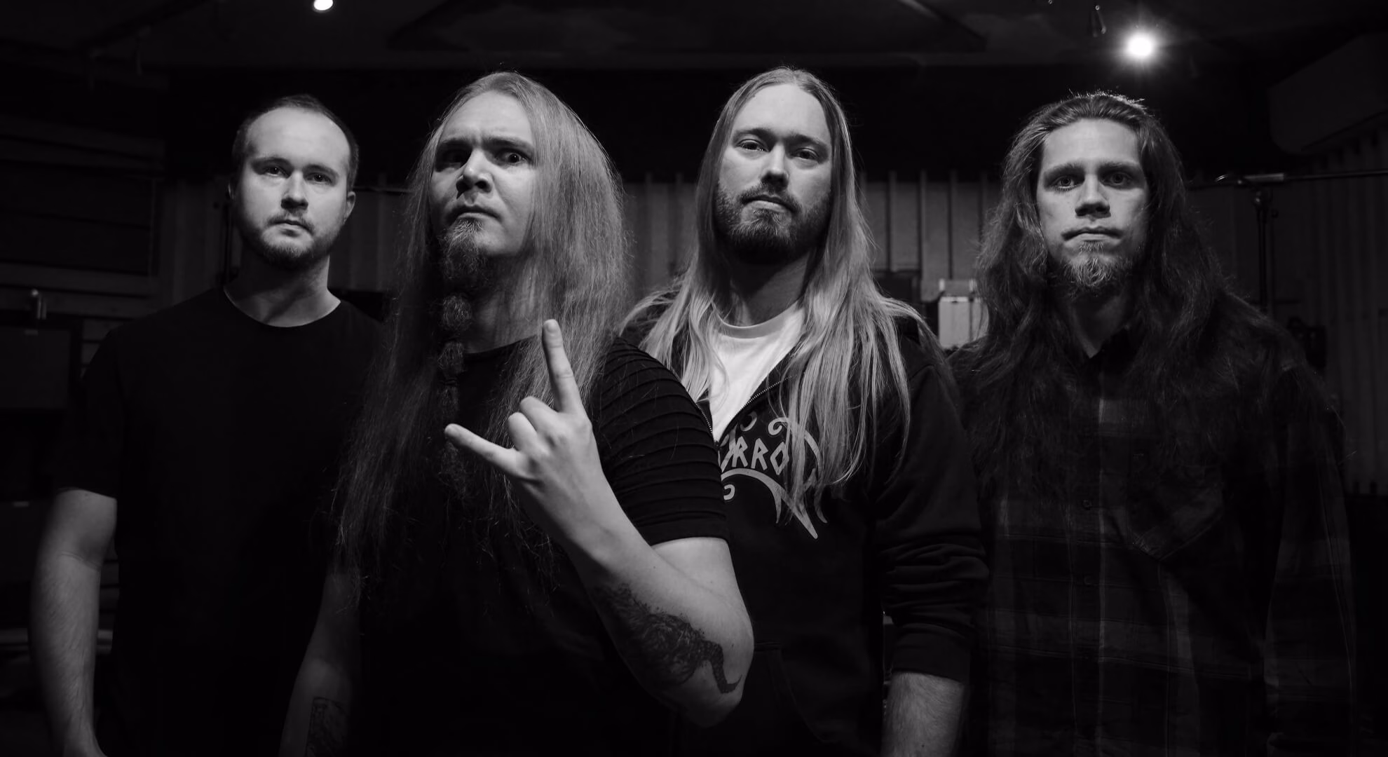 Four members of the metal band Inthraced pose in a studio. Three have long hair, and the central figure raises a hand gesture.