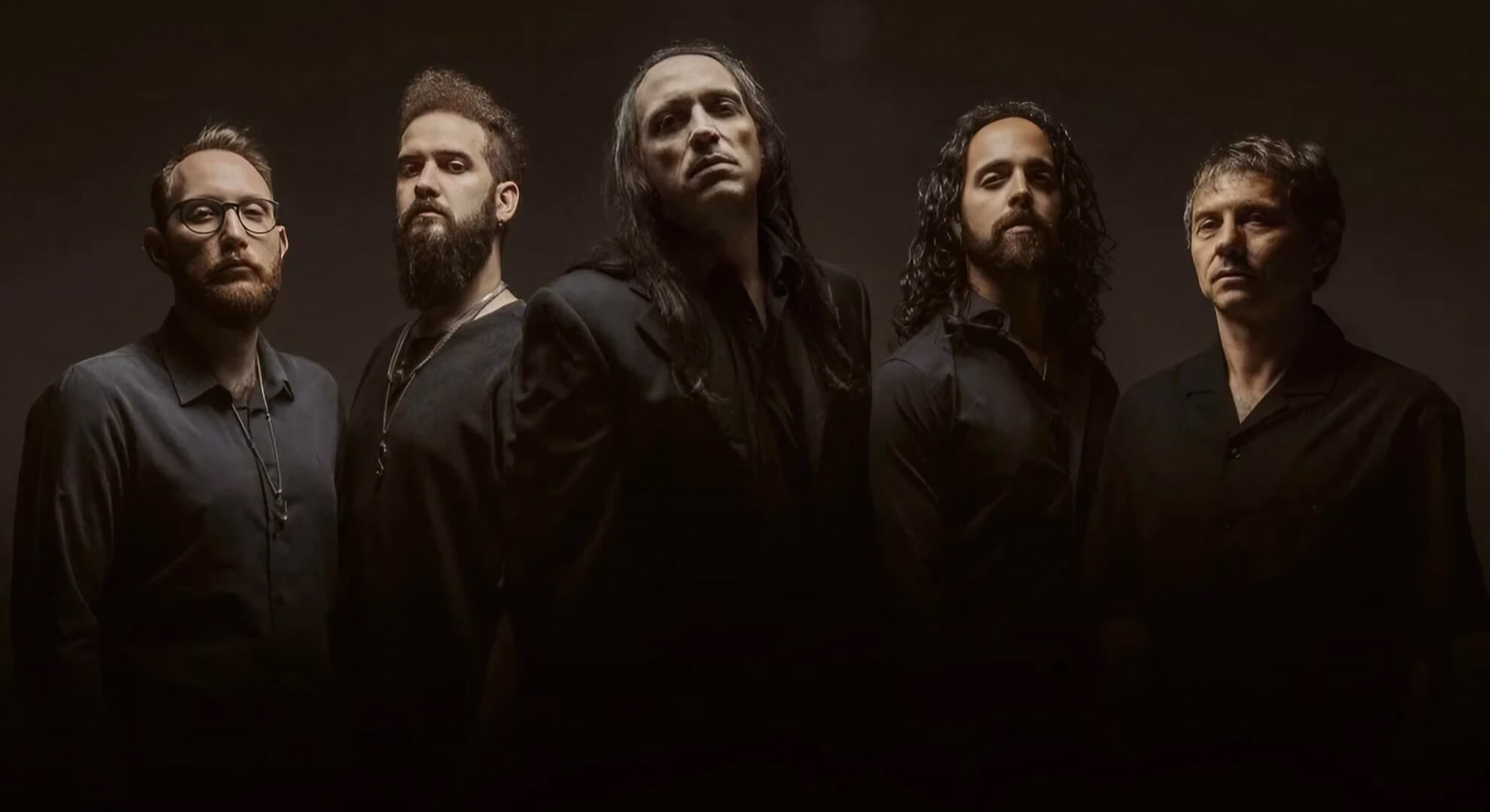 Five men in dark shirts stand in a line against a dark background, lit dramatically from the front and above.