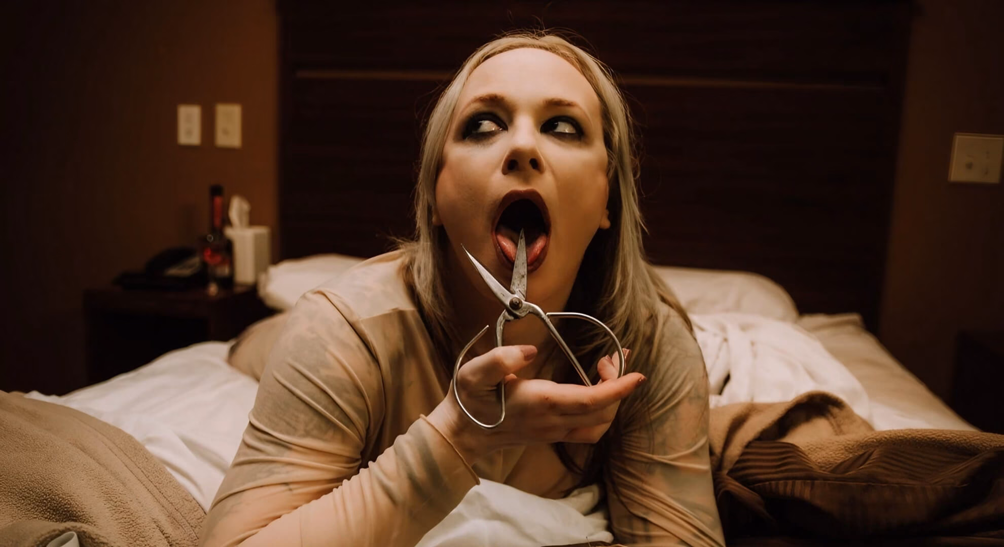 A blonde person with dark makeup lies on a bed, holding open scissors near their protruding tongue in a hotel room.