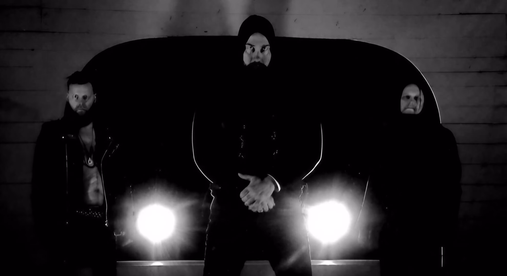 Three men in dark clothing stand in shadow, starkly backlit by two bright circular lights.