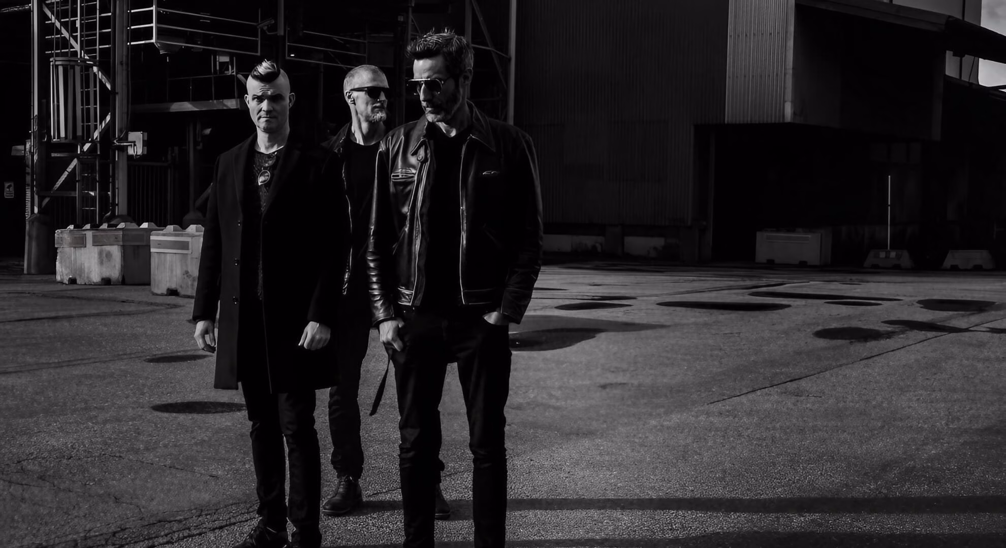Three band members in dark outerwear stand on sunlit concrete before a shadowy industrial structure in black and white.