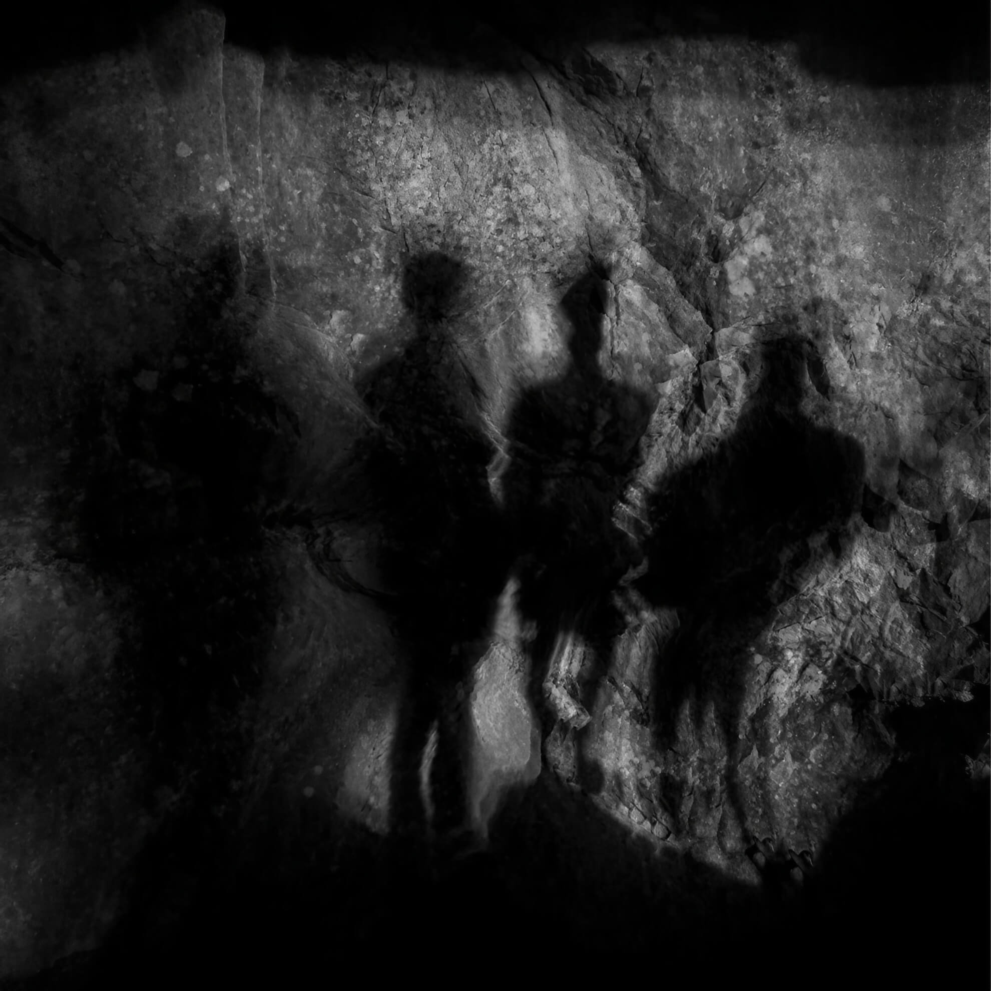 Four distorted, ghostly shadow silhouettes are cast against a dark, textured rock face in high-contrast grayscale.