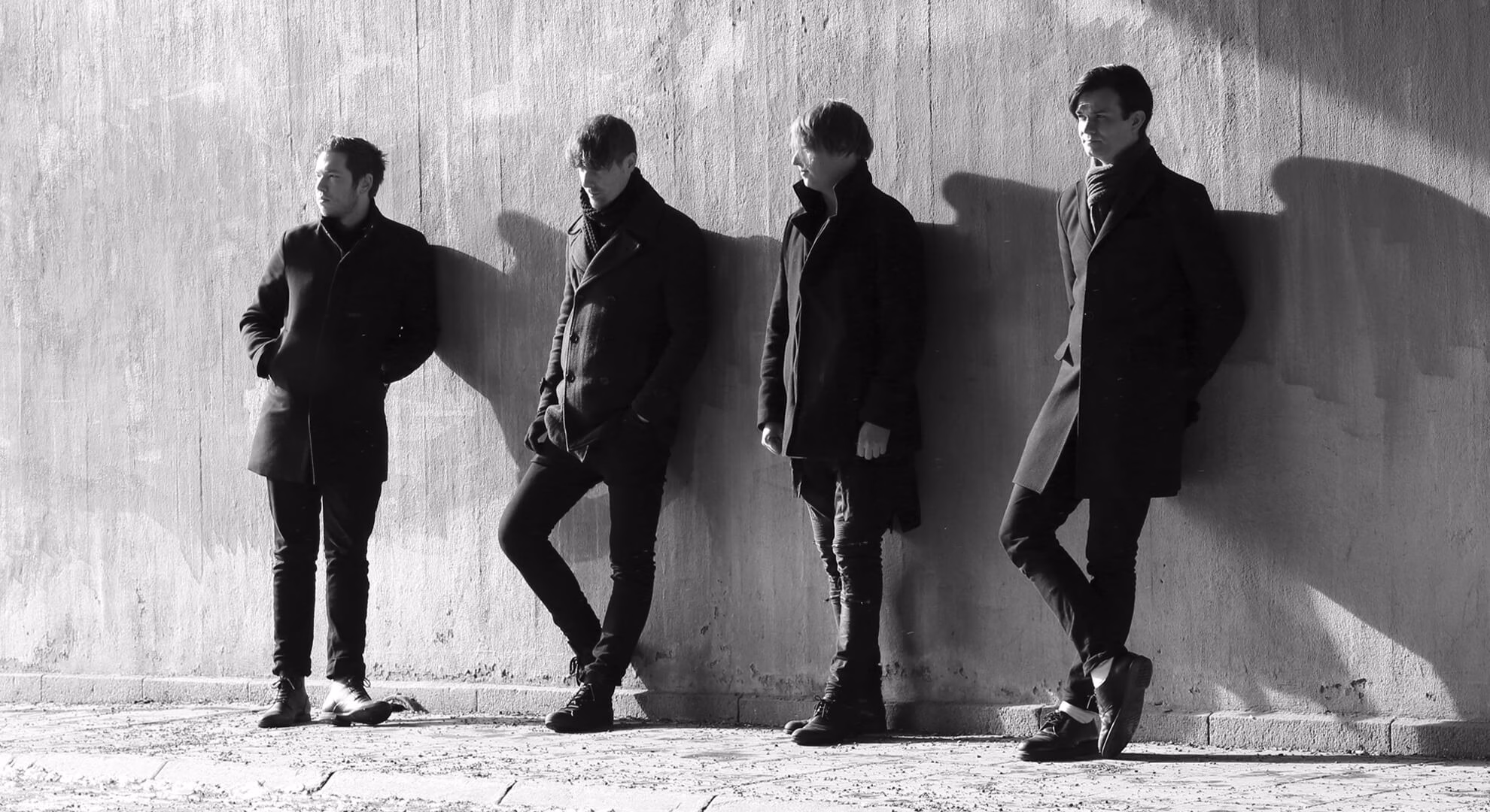 Four men in dark coats and sunglasses stand on concrete before a shadowy industrial structure in black and white.