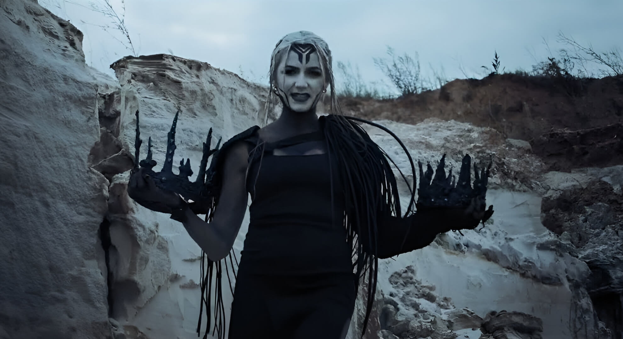 A vocalist with white makeup and black facial lines stands in a quarry, holding jagged black crowns in each hand under soft natural light.