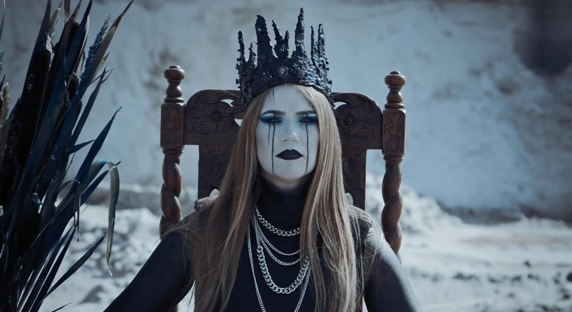 A vocalist sits on a wooden throne in a white, sandy quarry, wearing a jagged black crown and heavy silver chains, with black tears painted on her white face.