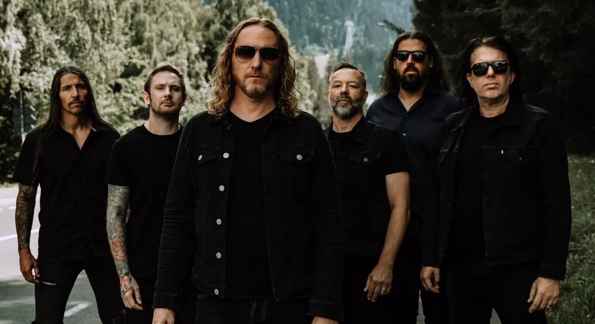 Six band members in dark clothing stand on a tree-lined road; the central figure wears sunglasses and a black denim jacket.