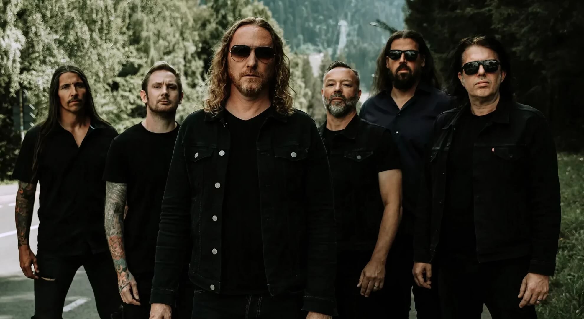 Six band members in dark clothing stand on a tree-lined road; the central figure wears sunglasses and a black denim jacket.
