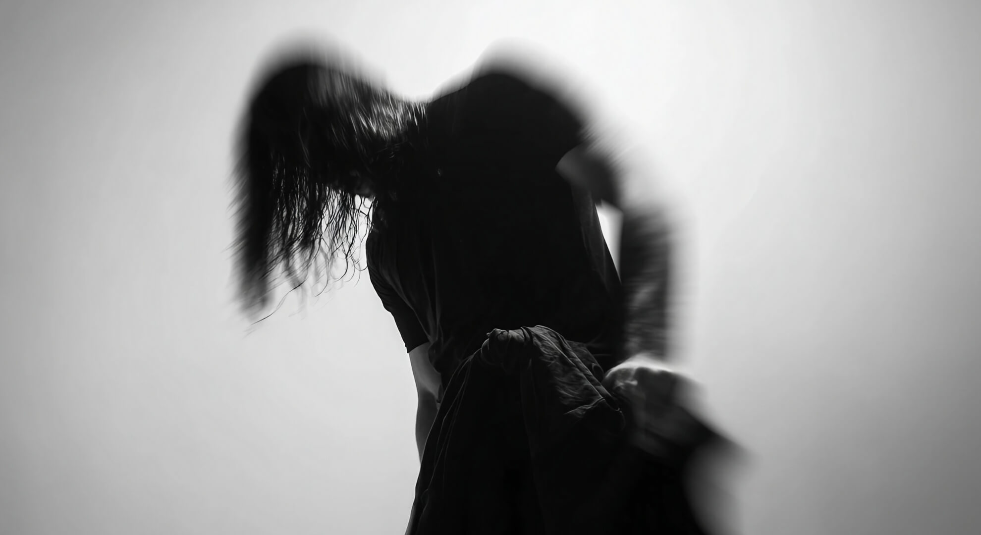 Black and white photograph showing a person with long dark hair in motion blur, head tilted back with hand near face, creating a dramatic sense of movement.