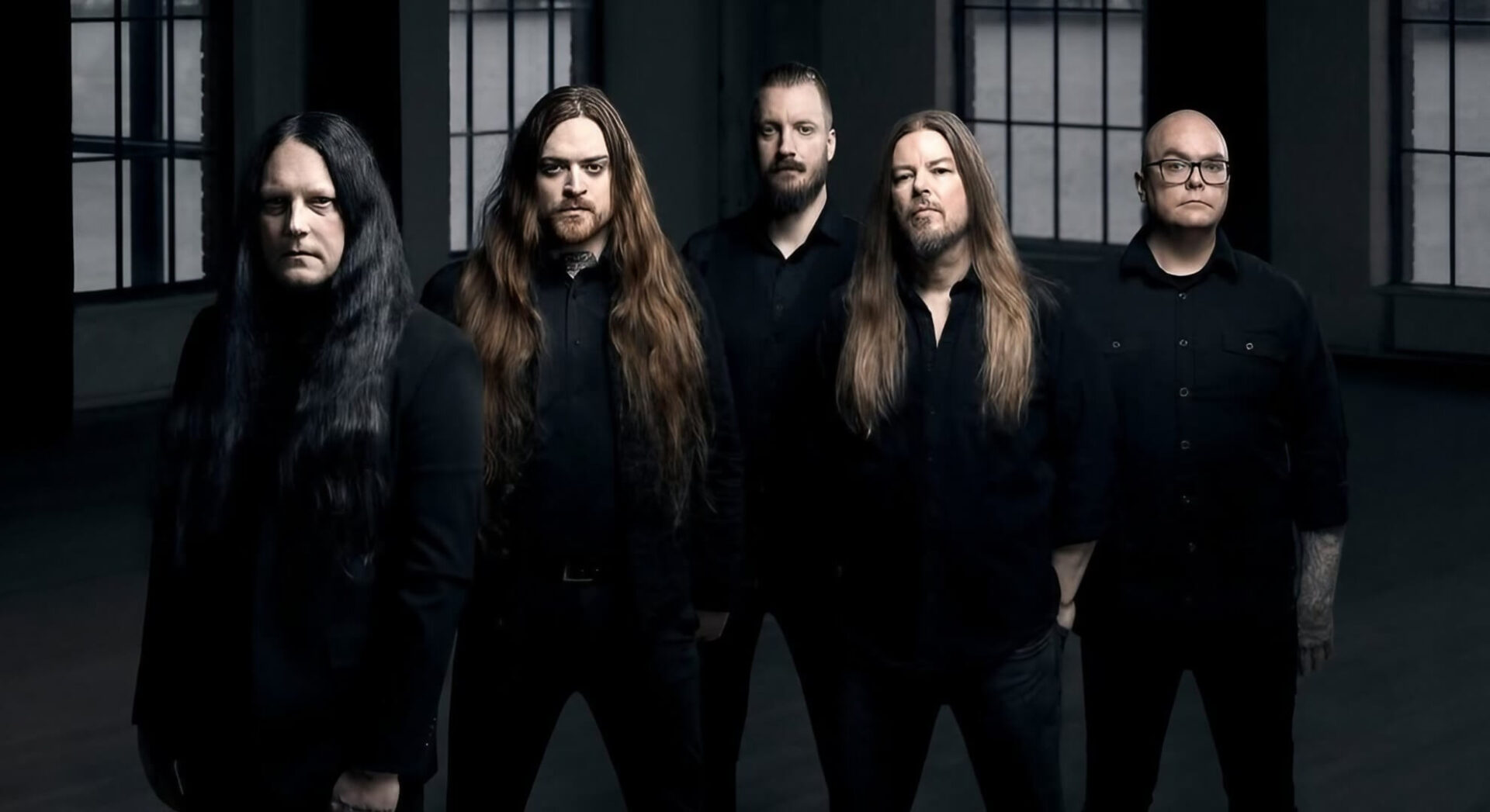 A somber group portrait of five men in black attire standing in a dimly lit room with large industrial windows.
