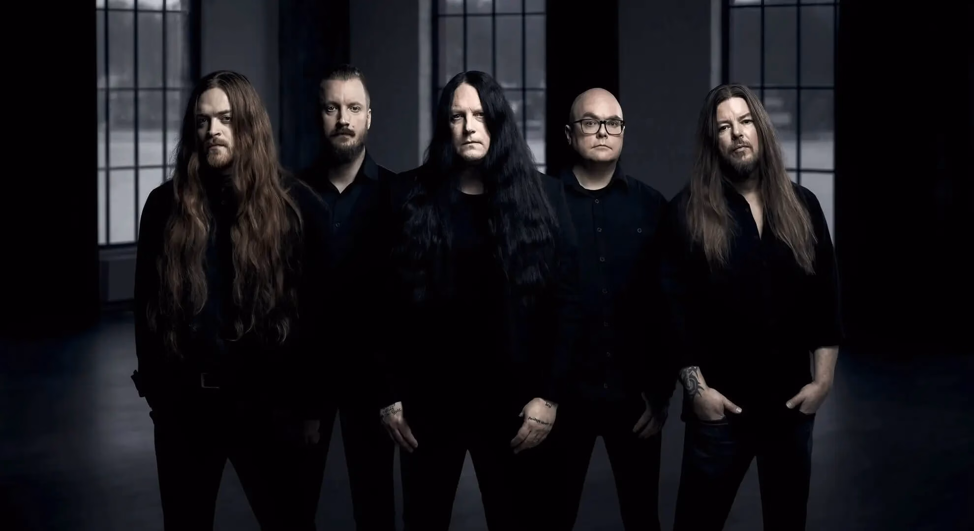 Five men in black clothing stand in a row in a dimly lit room with tall windows in the background.