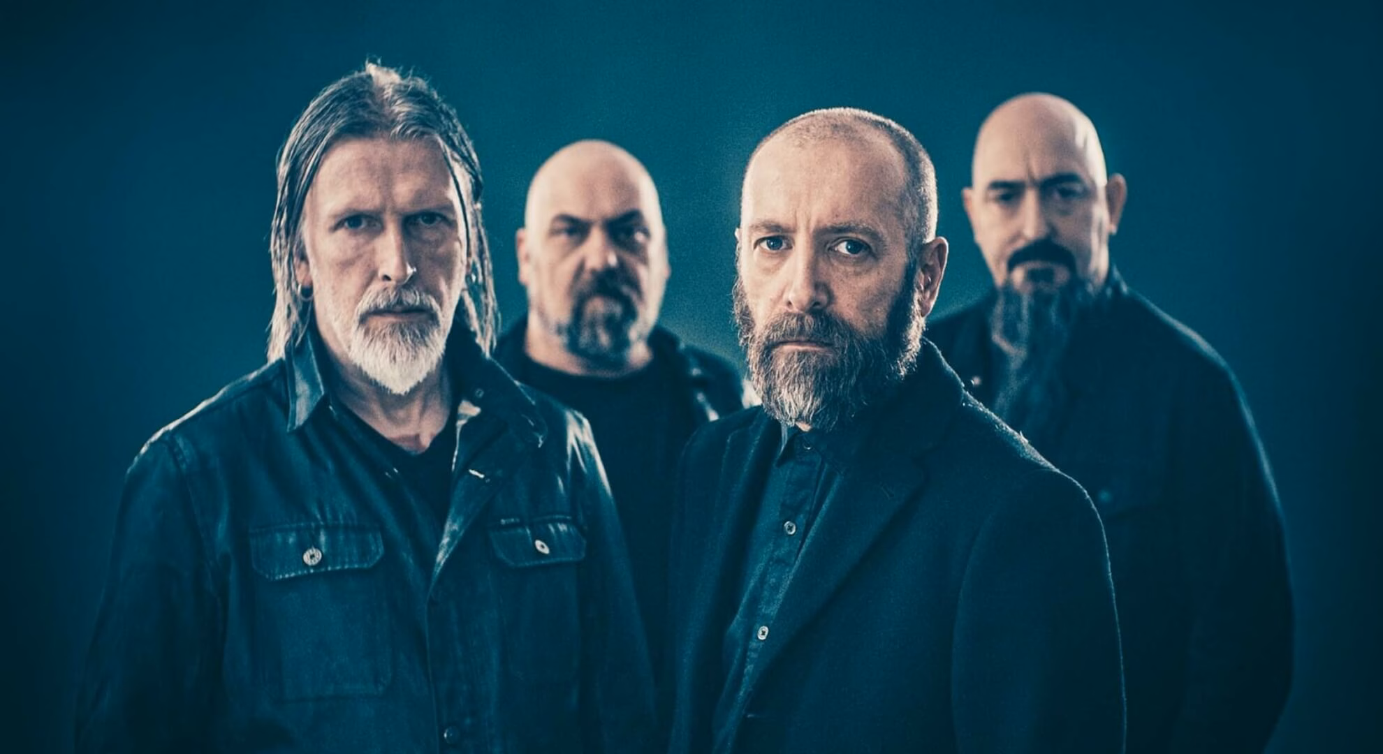 Paradise Lost: Surveying the Terrain of Melancholy in ‘Salvation’ A desaturated, medium shot of Paradise Lost’s four members standing against a stark, textured grey backdrop. They are dressed in dark overcoats, gazing solemnly forward, flanked by deep shadows that obscure the edges of the frame.