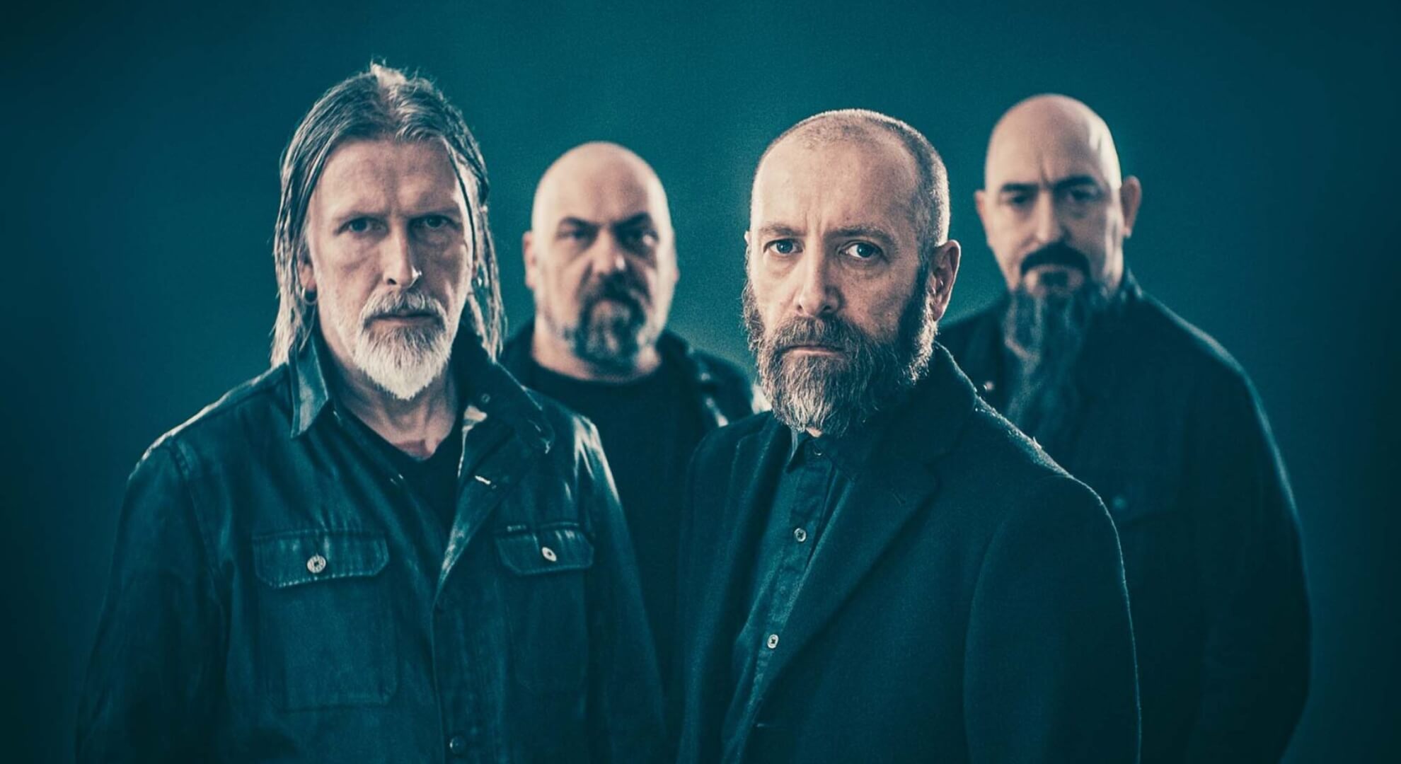 Paradise Lost: Surveying the Terrain of Melancholy in ‘Salvation’ A desaturated, medium shot of Paradise Lost’s four members standing against a stark, textured grey backdrop. They are dressed in dark overcoats, gazing solemnly forward, flanked by deep shadows that obscure the edges of the frame.