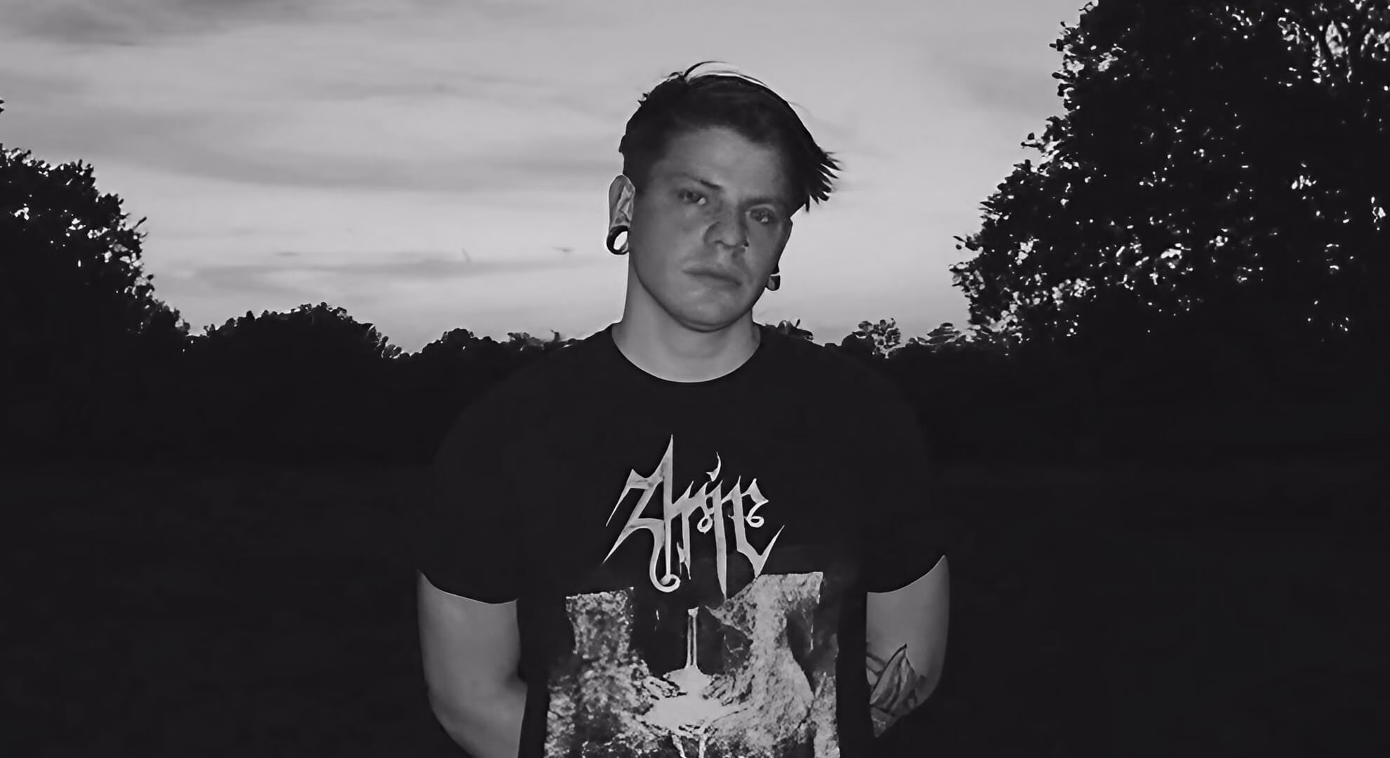 Wyatt Sharp stands outdoors at dusk in black and white, wearing ear gauges and a dark graphic t-shirt against silhouetted trees.