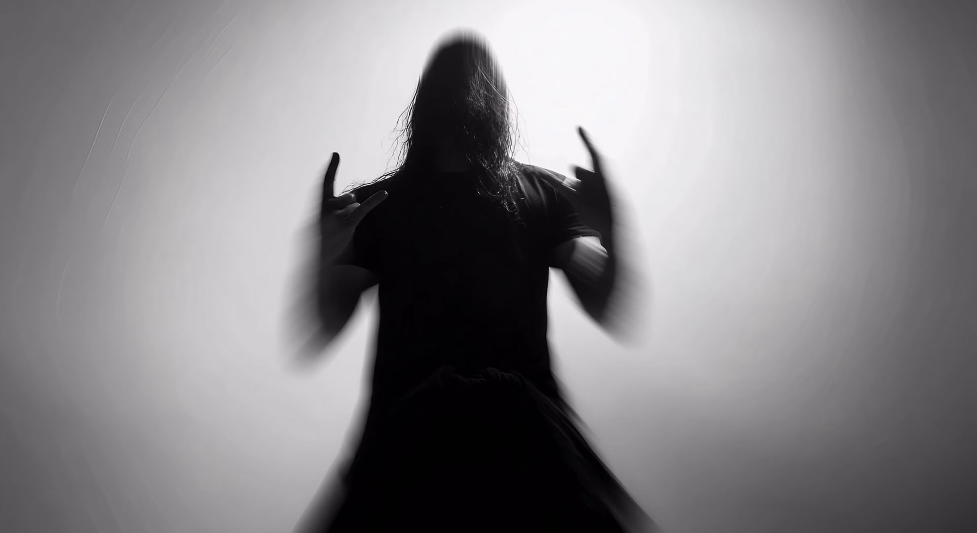 Black and white photograph of a silhouetted figure with long dark hair, arms raised with hands making the metal horns gesture.