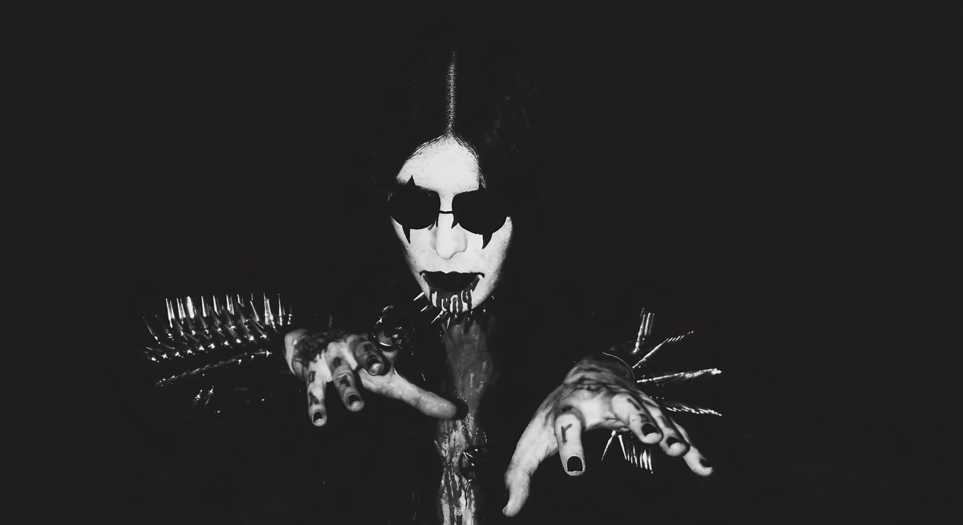 A black-and-white shot of a musician in corpse paint and spiked gauntlets, reaching toward the camera with clawed hands.