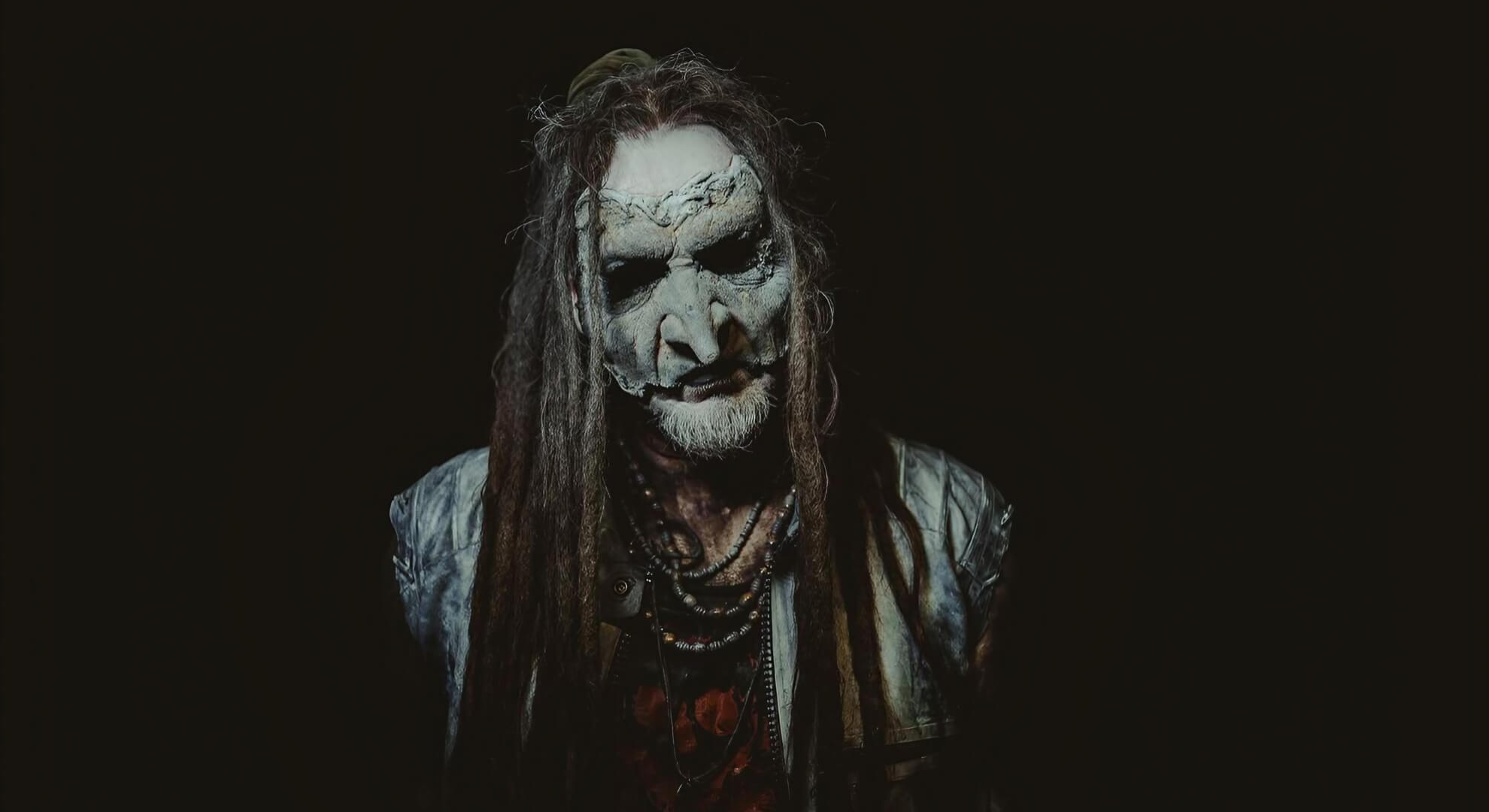 A portrait of Mortiis in his signature prosthetic goblin mask and dreadlocks, set against a dark, atmospheric backdrop.