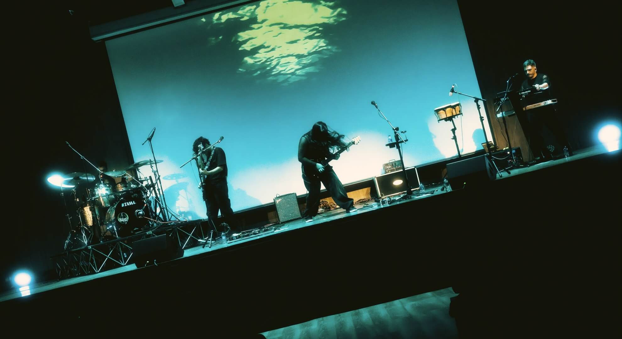 Argovia performing on stage with cool blue-teal lighting and water patterns on projection screen.