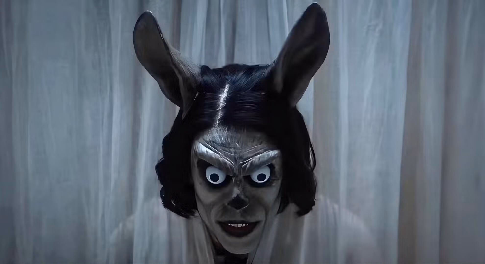 &lsquo;Hokum&rsquo; (2026) film still: a figure in a grey horned mask with googly eyes affixed over the eye sockets, dark hair, white curtains behind.
