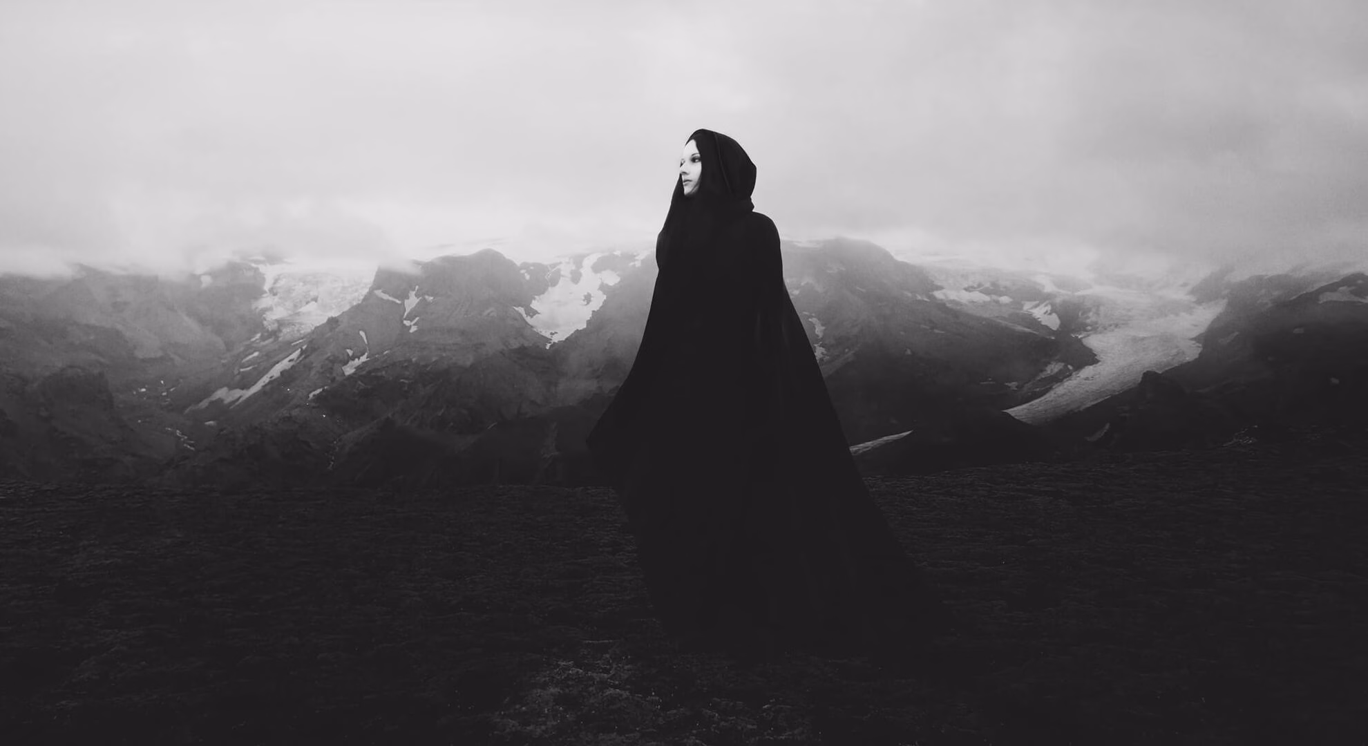 Heike Langhans in a black hooded cape standing against a fog-covered Icelandic mountain range.