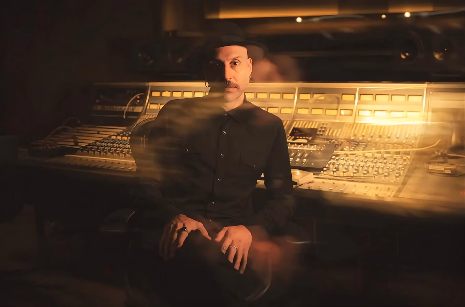 Jaime Gomez Arellano seated at a mixing console in amber studio light, hands resting, facing the camera.