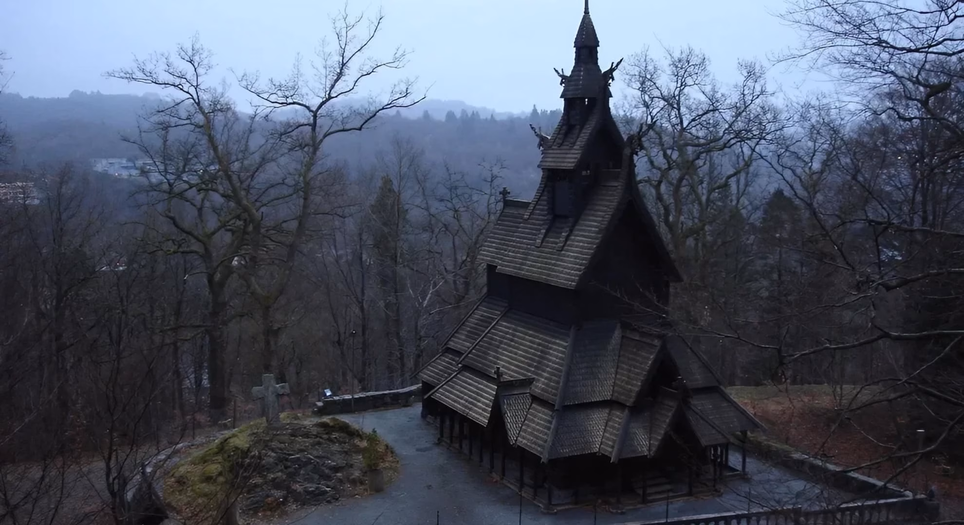 Fantoft Stave Church at dusk, viewed through bare winter trees with a stone cross in front.