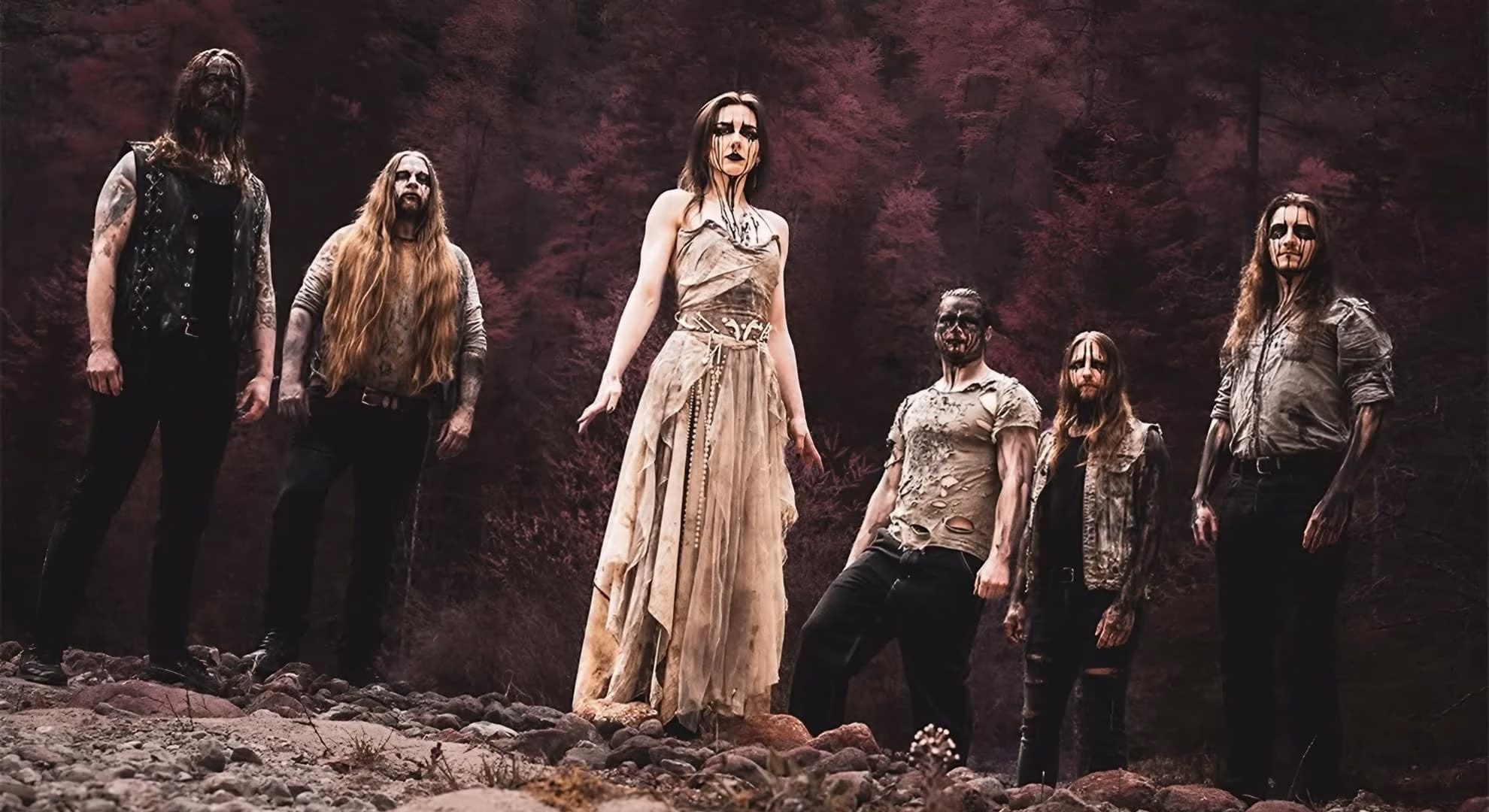 Firtan Turn the Annihilating Light Inward on ‘Helios’ Six corpse-painted Firtan figures stand across rocky ground before a dark red forest, the central figure in a pale gown.