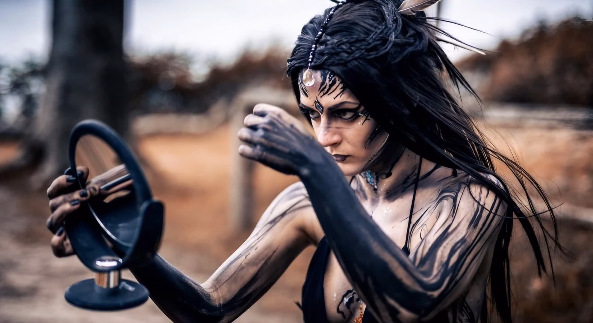 M’era Luna Turns 26 on the Hildesheim Airfield Body-painted performer in dark ritual makeup and feathered headdress holds a small mirror outdoors.