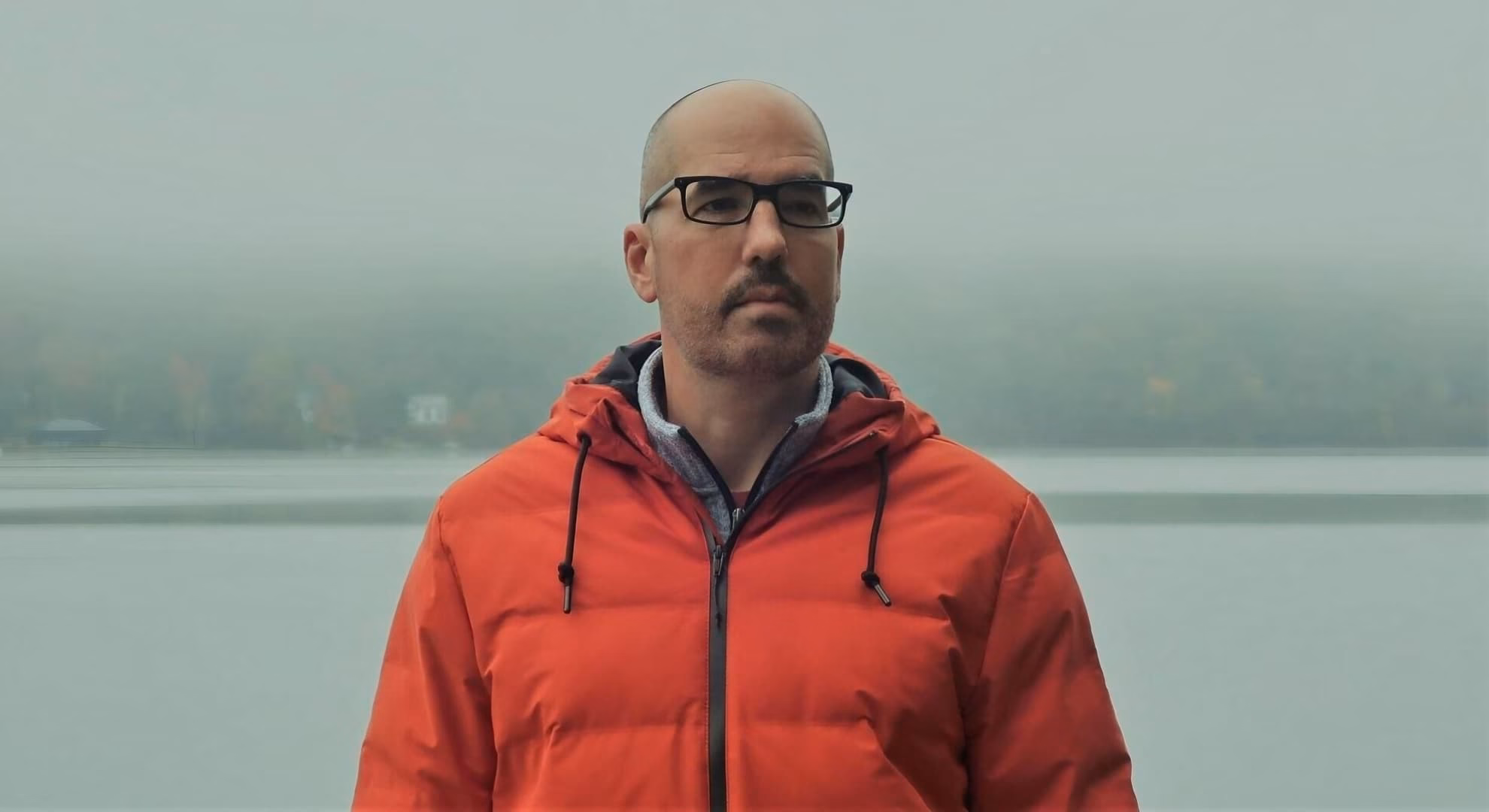 Riley Sager, author of ‘The Unknown,’ stands before a still lake and its shoreline.